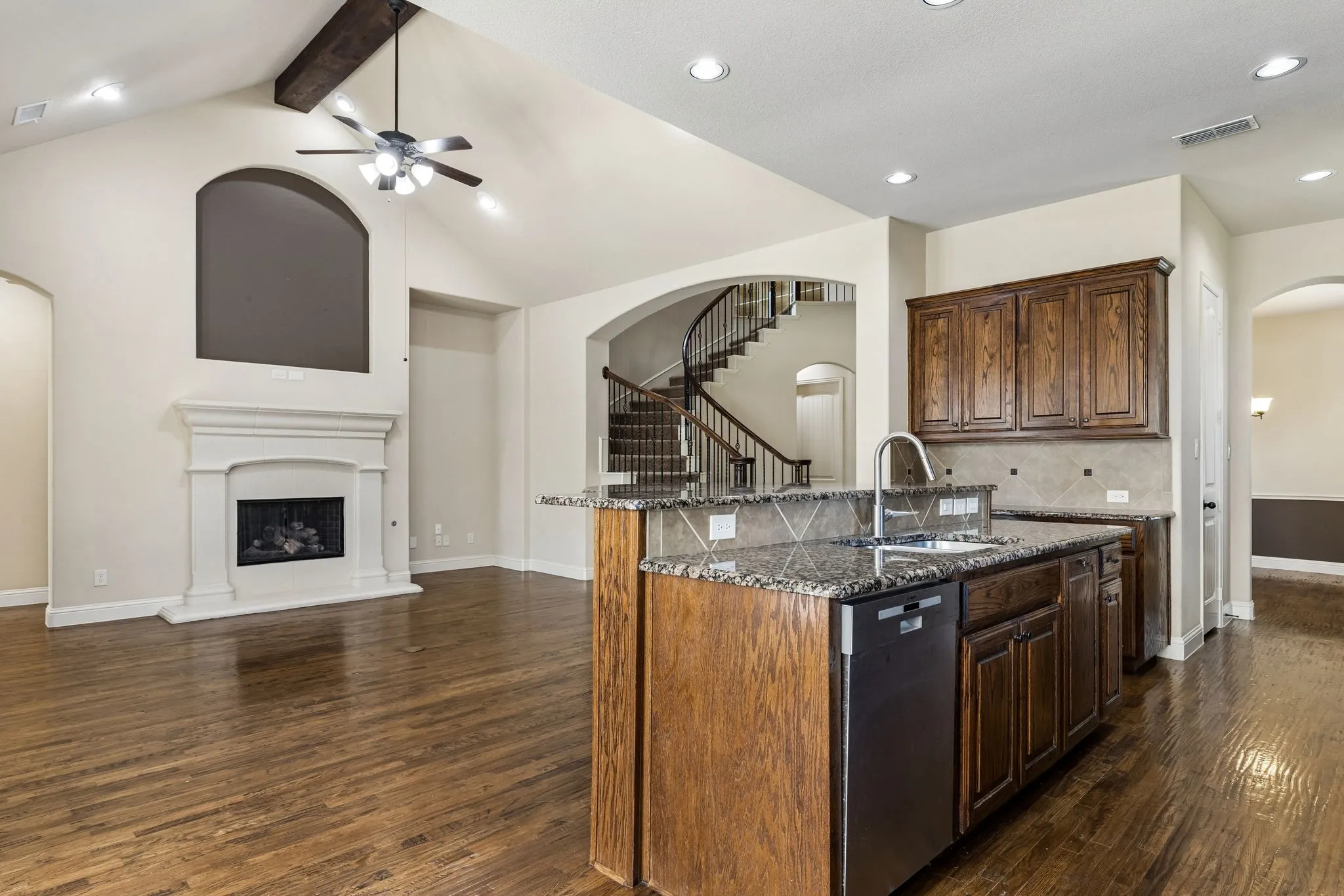 Vaulted ceilings with exposed timber beams, wood-finish flooring, masonry fireplace with decorative surround, kitchen island with stone-surface countertop, and curved staircase with wrought-iron railing