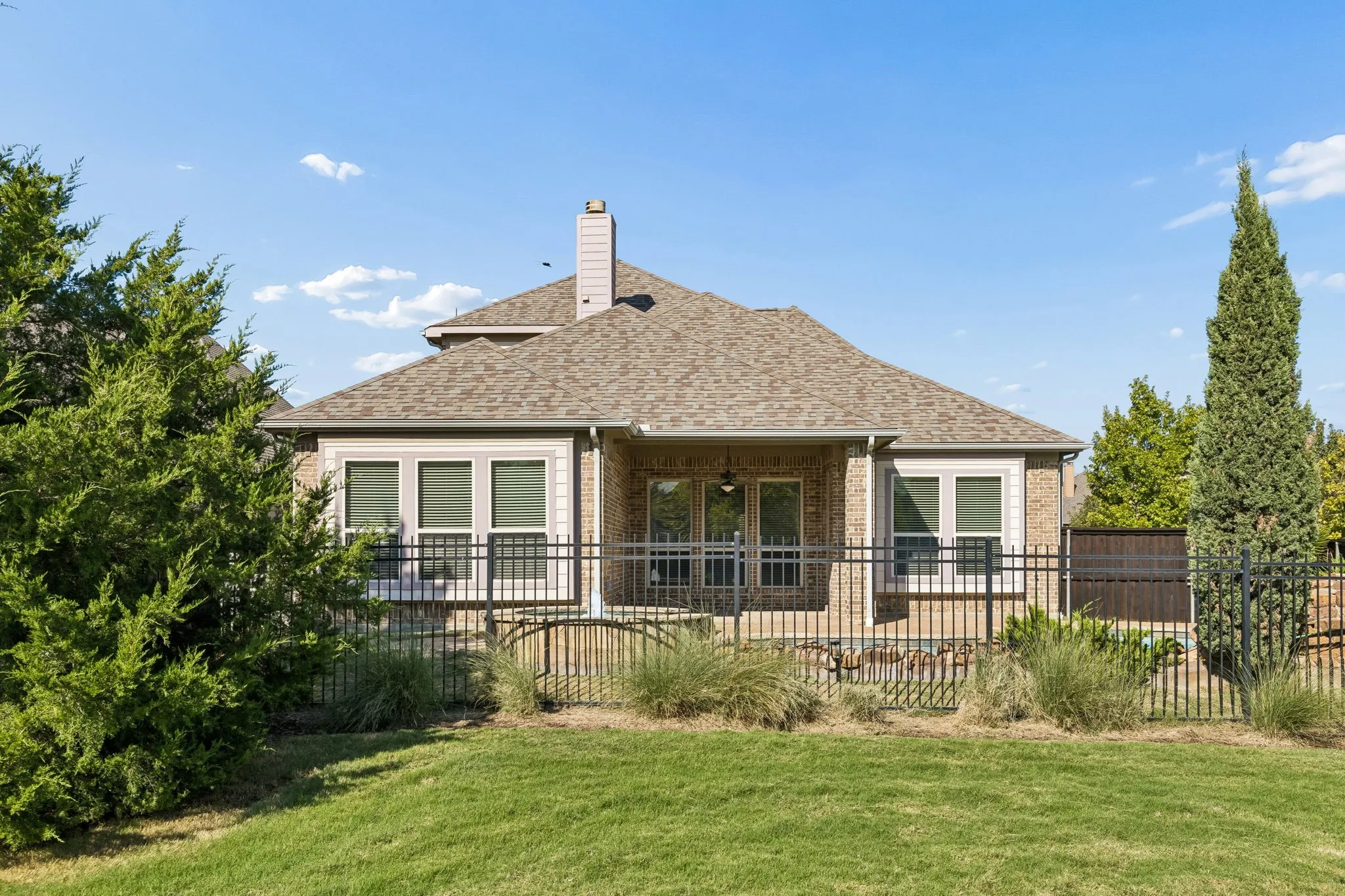 Rear elevation featuring a brick exterior, gabled roofline, centralized masonry chimney, covered patio, and perimeter metal fencing