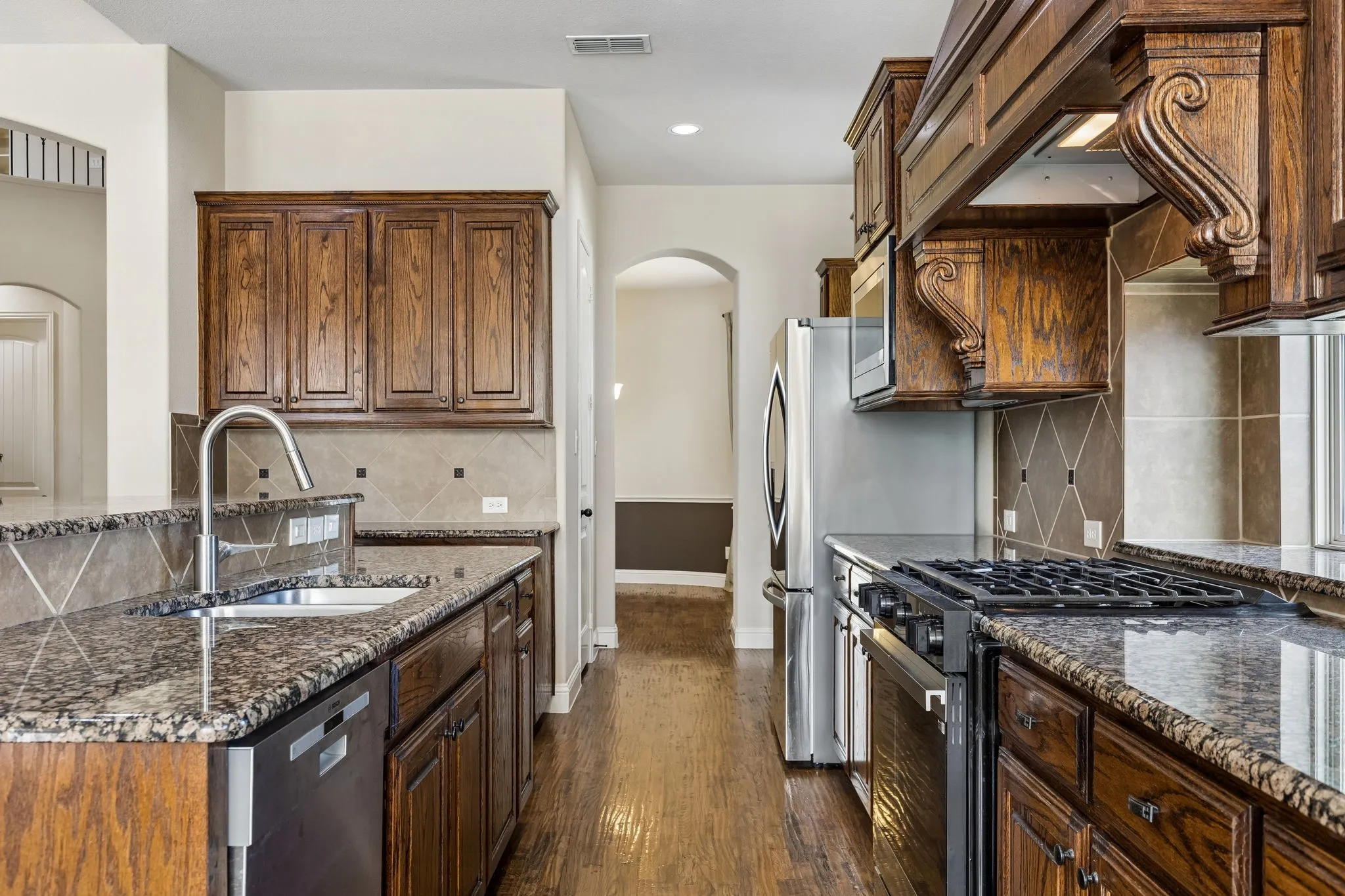 Kitchen featuring wood-finish cabinetry, stone countertops, a decorative range hood, stainless steel appliances, and diagonal tile backsplash