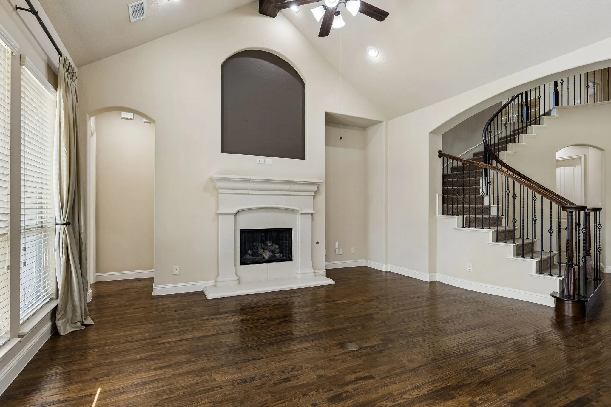 Vaulted ceiling, wood-finish flooring, central fireplace with a classic mantel, and a curved staircase featuring wrought-iron balusters
