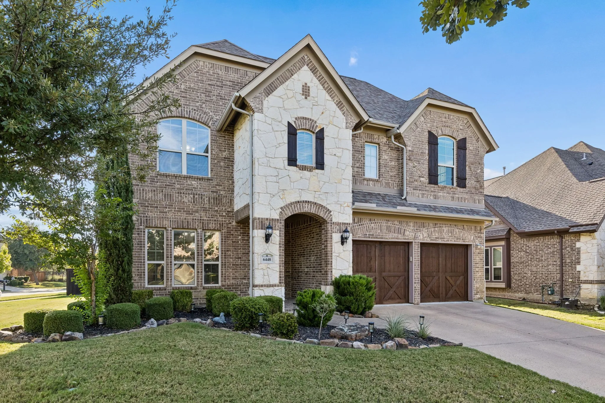Two-story brick and stone veneer exterior featuring a covered arched entryway, decorative dark wood-finish carriage-style garage doors, dark shutters, and a multi-gabled roofline