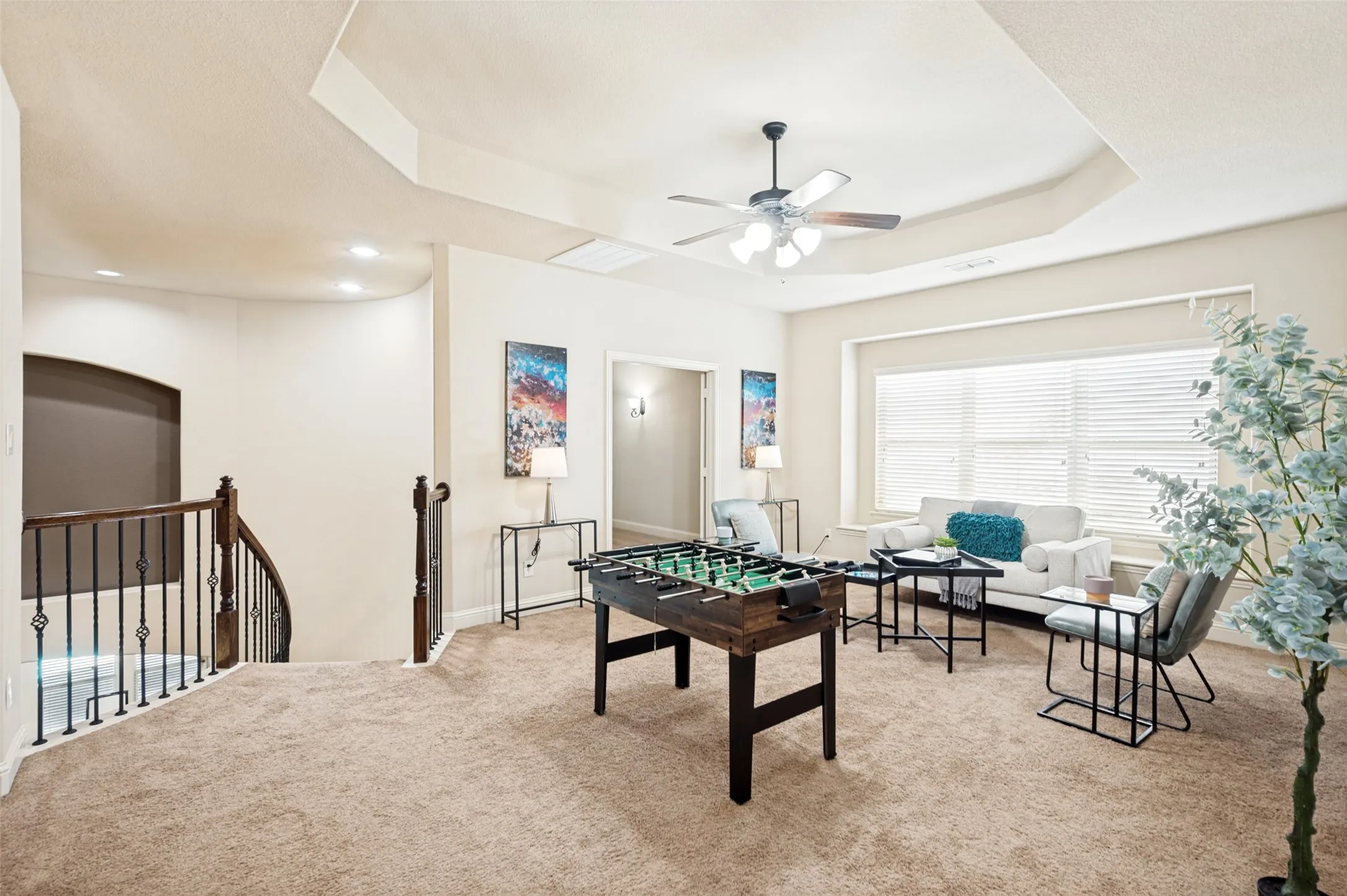 Tray ceiling with a central ceiling fan, windowed wall with horizontal blinds, and a staircase featuring a wooden handrail and metal balusters