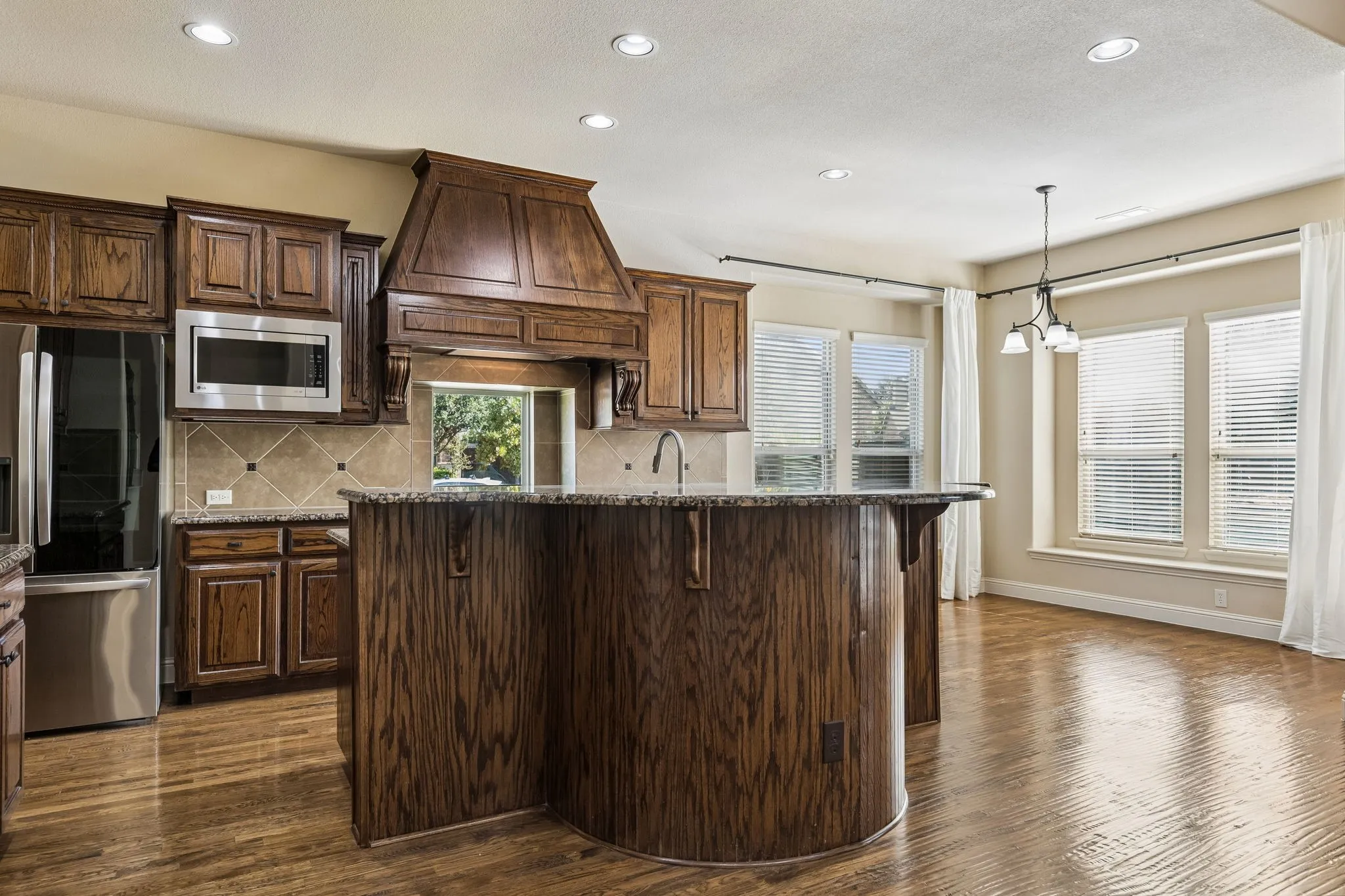 Kitchen featuring custom wood cabinetry, a curved island with stone countertops, a decorative wood range hood, tile backsplash, and wood-finish flooring