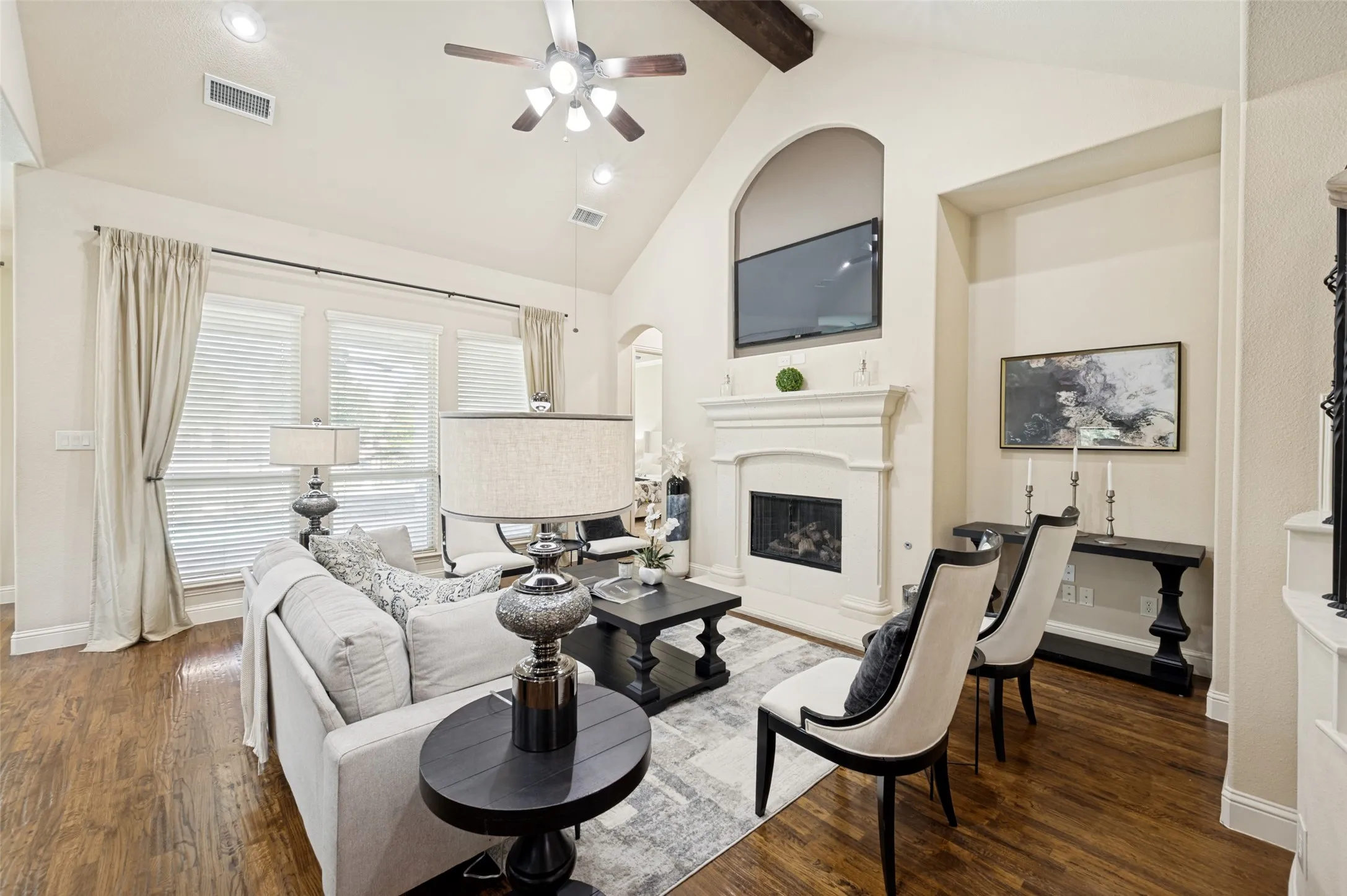 Vaulted ceiling featuring a decorative wood beam, stone-surround fireplace with a recessed alcove, large windows with multi-pane configuration, and wood-finish flooring
