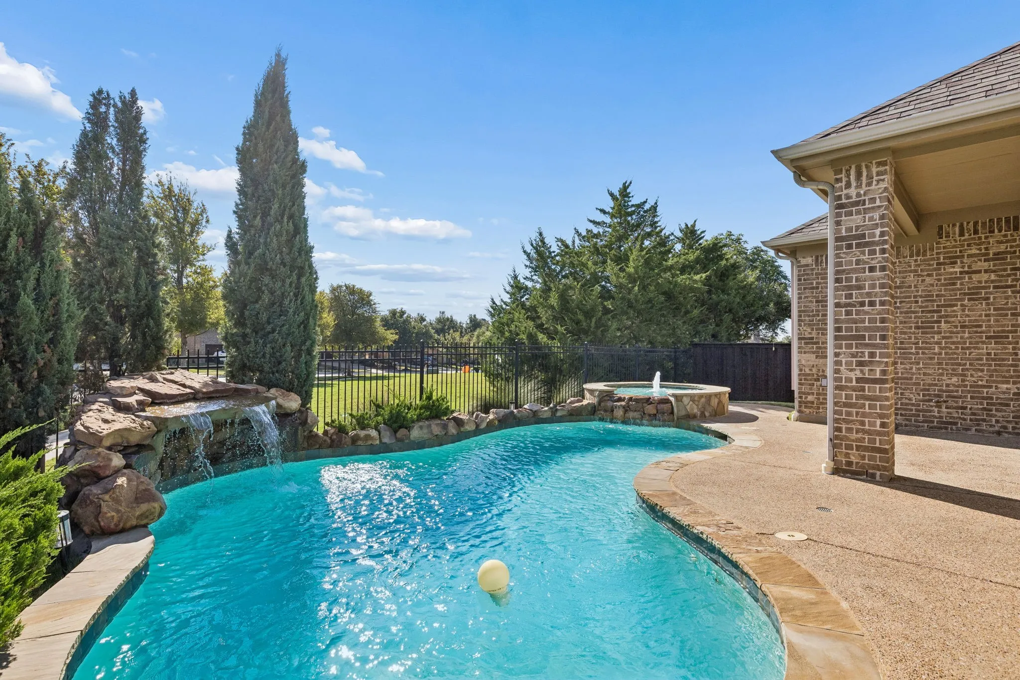 Outdoor pool area featuring a natural rock waterfall, elevated spill-over spa, textured concrete decking, and brick exterior finish