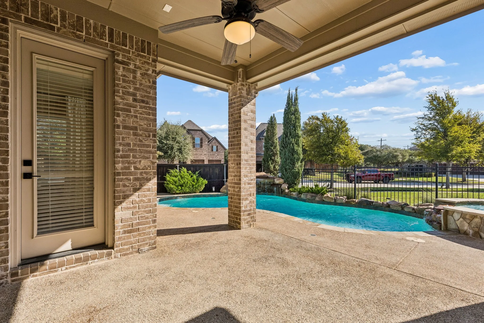 Covered patio featuring a brick exterior, recessed ceiling, and an integrated overhead fan, overlooking an in-ground pool with a stone-accented spa and perimeter metal fencing