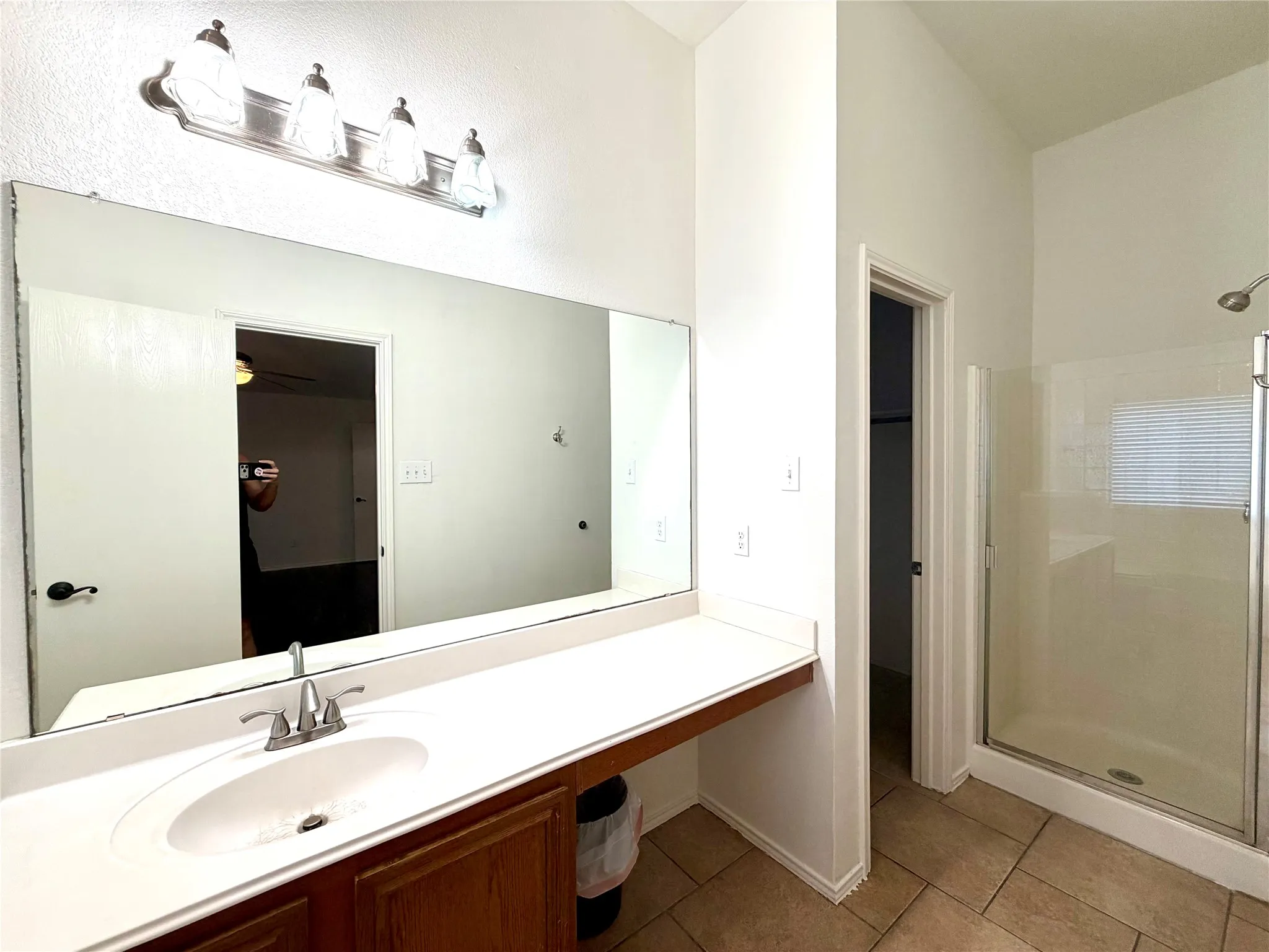 Bathroom featuring a single-sink vanity with a solid-surface countertop, a full-width wall mirror, a four-light fixture, and a glass-enclosed shower stall with tile flooring