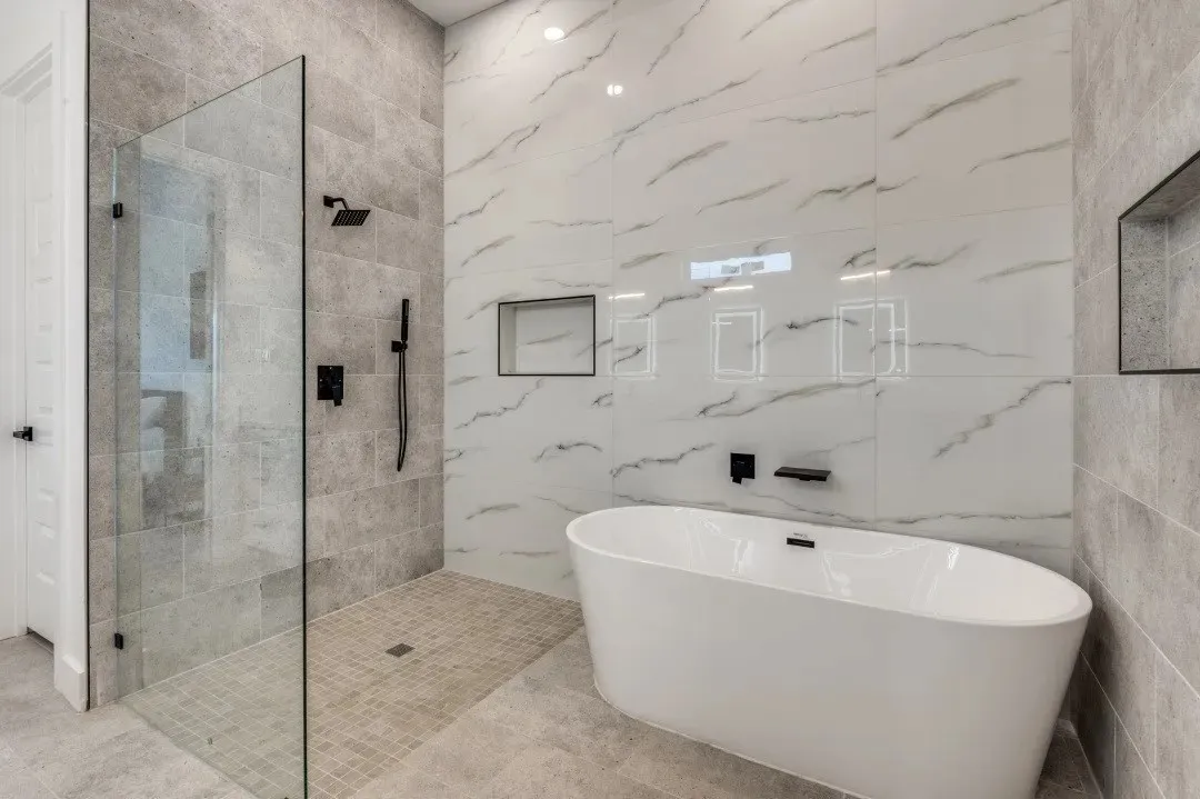 Freestanding soaking tub with matte black fixtures, walk-in shower featuring a frameless glass enclosure, large-format stone-finish wall tile, and recessed wall niches