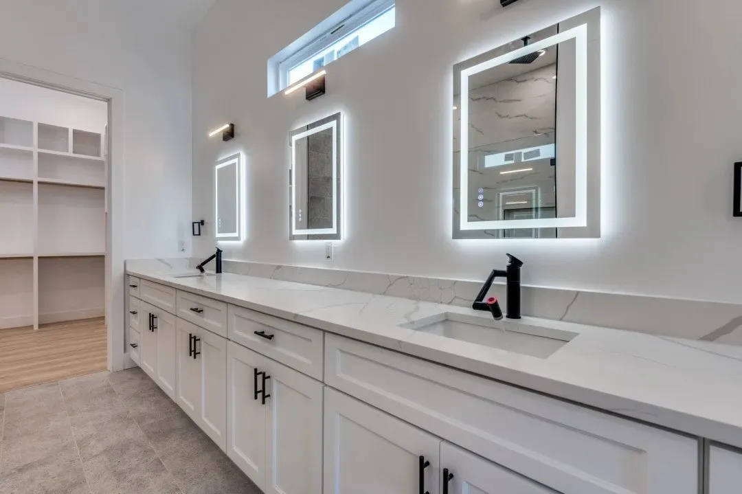 Dual-sink vanity with stone-surfaced countertop, white shaker-style cabinetry, matte black hardware, integrated backlit mirrors, and wall-mounted light fixtures