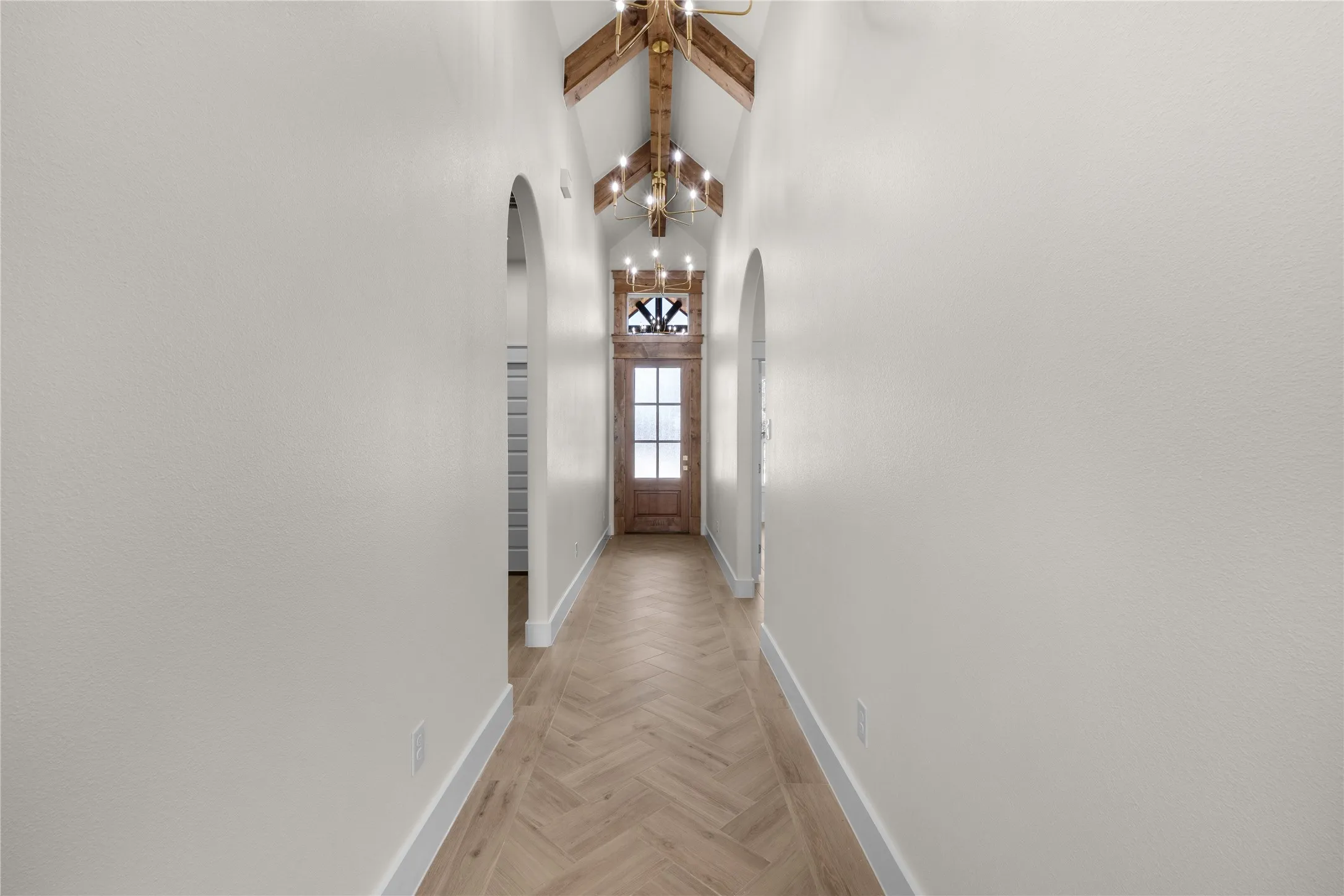 Vaulted ceiling featuring exposed wood beams, herringbone-patterned wood-finish flooring, arched doorways, and a wood-framed entry door with a decorative transom window