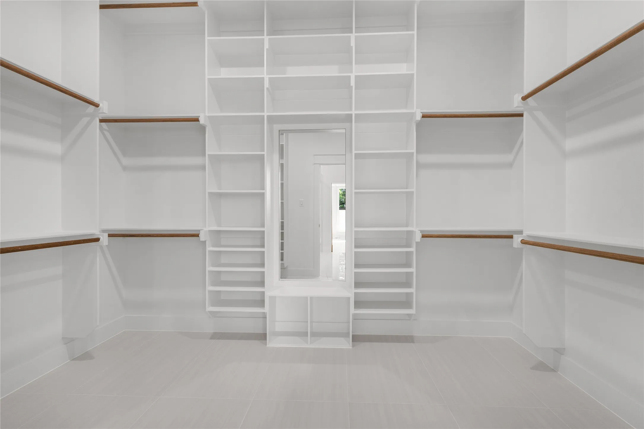 Custom walk-in closet featuring floor-to-ceiling white shelving, integrated hanging rods with wood finishes, a central built-in vanity mirror, and ceramic tile flooring