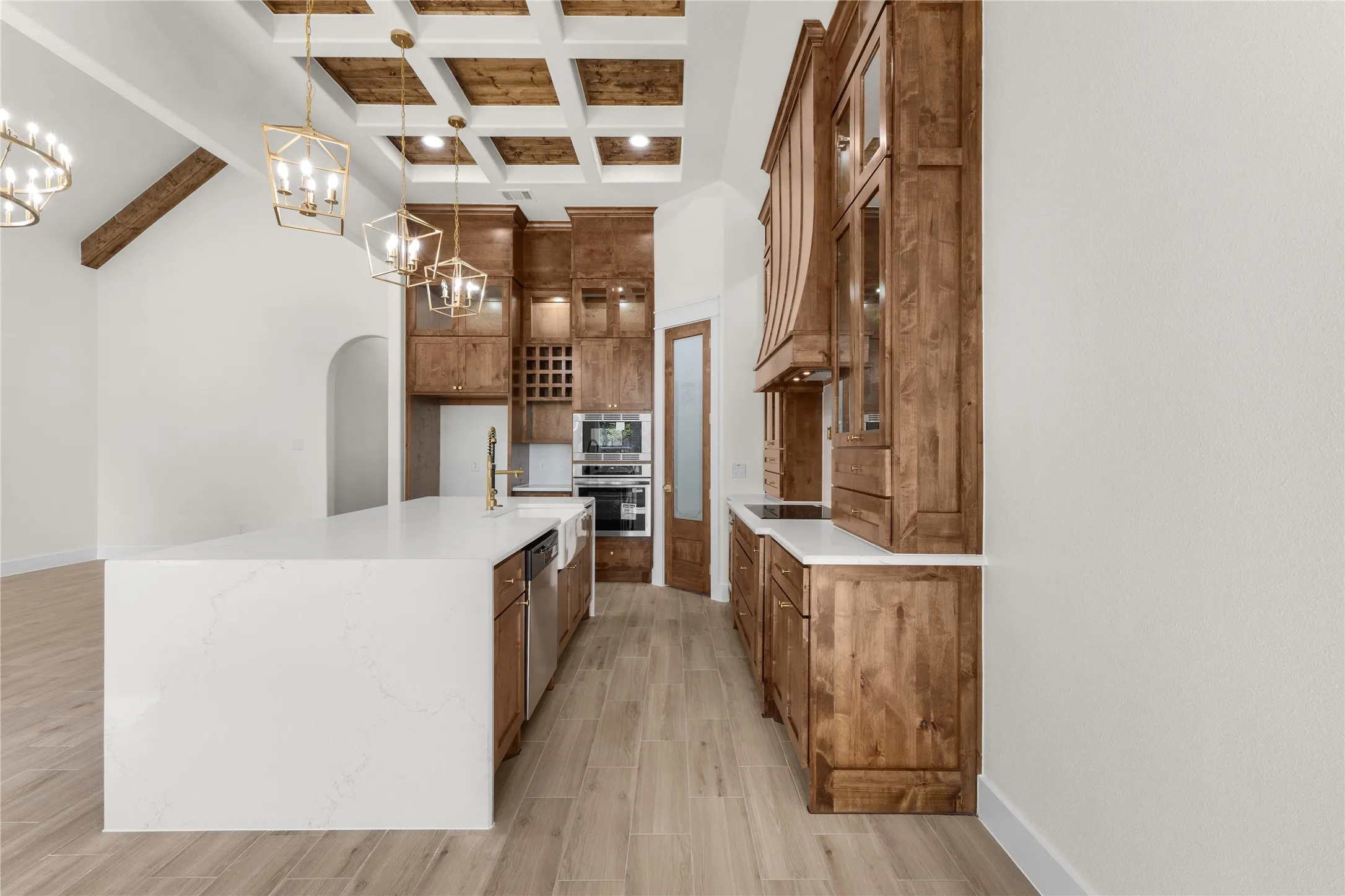 Kitchen featuring custom stained-wood cabinetry, a large waterfall island with quartz countertops, a coffered ceiling with wood-finish inlays, integrated stainless steel double ovens, and wood-finish flooring