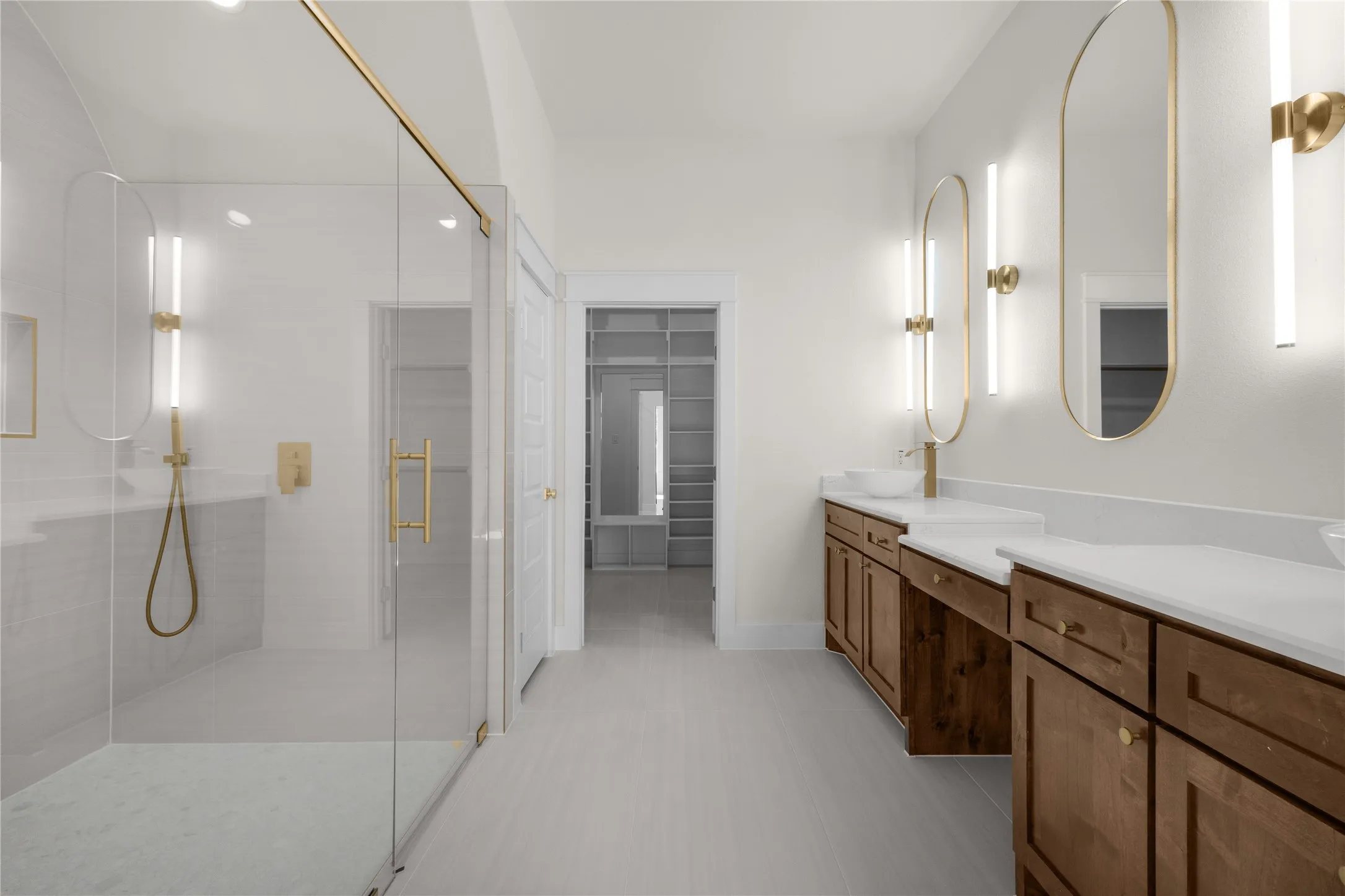 Glass-enclosed walk-in shower with gold-finish hardware, dual vanity with solid-surface countertops, vessel sinks, oval-framed wall mirrors, and integrated shelving in the dressing area