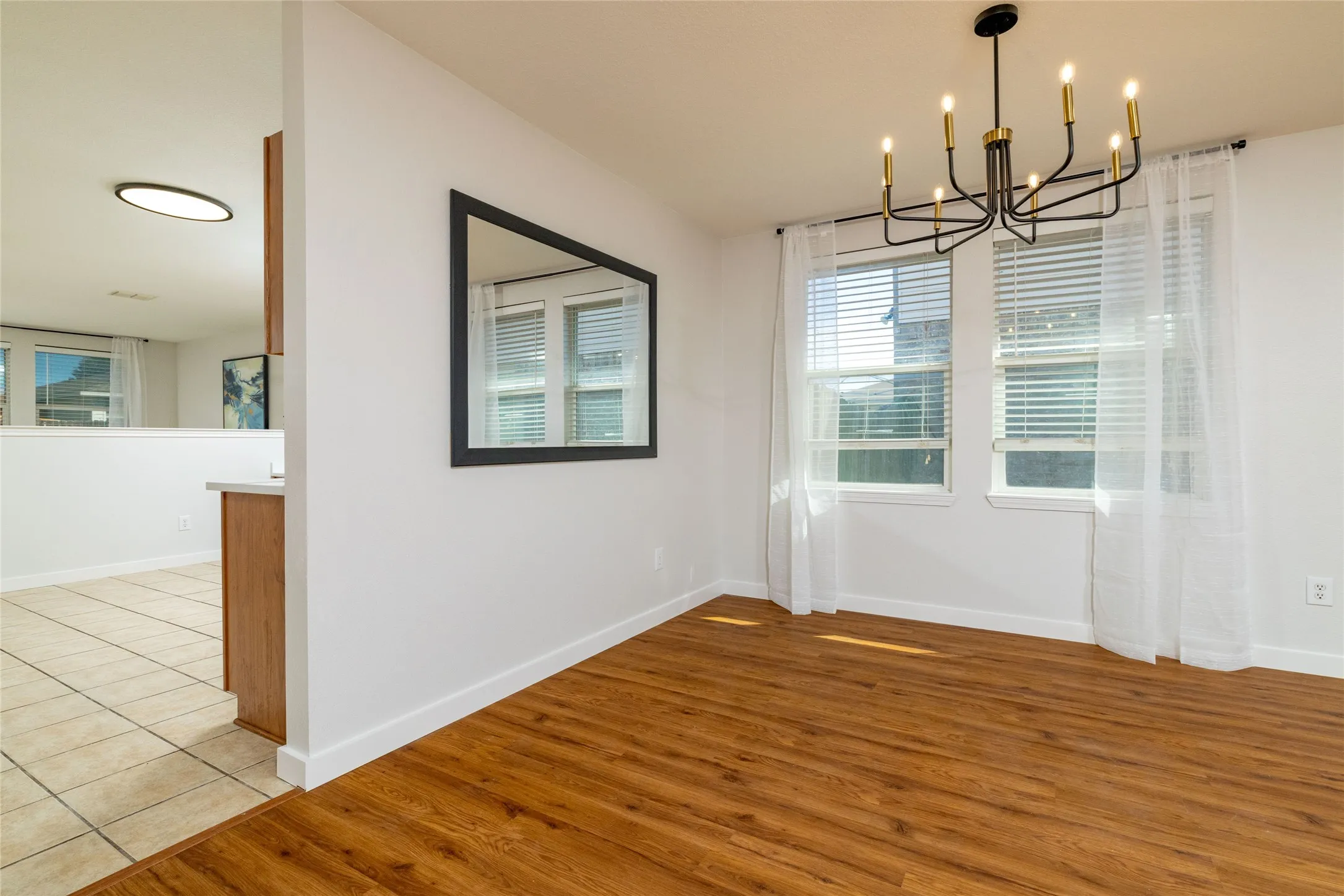 Wood-finish flooring, matte black multi-arm chandelier, dual windows with horizontal blinds, and a large wall-mounted mirror
