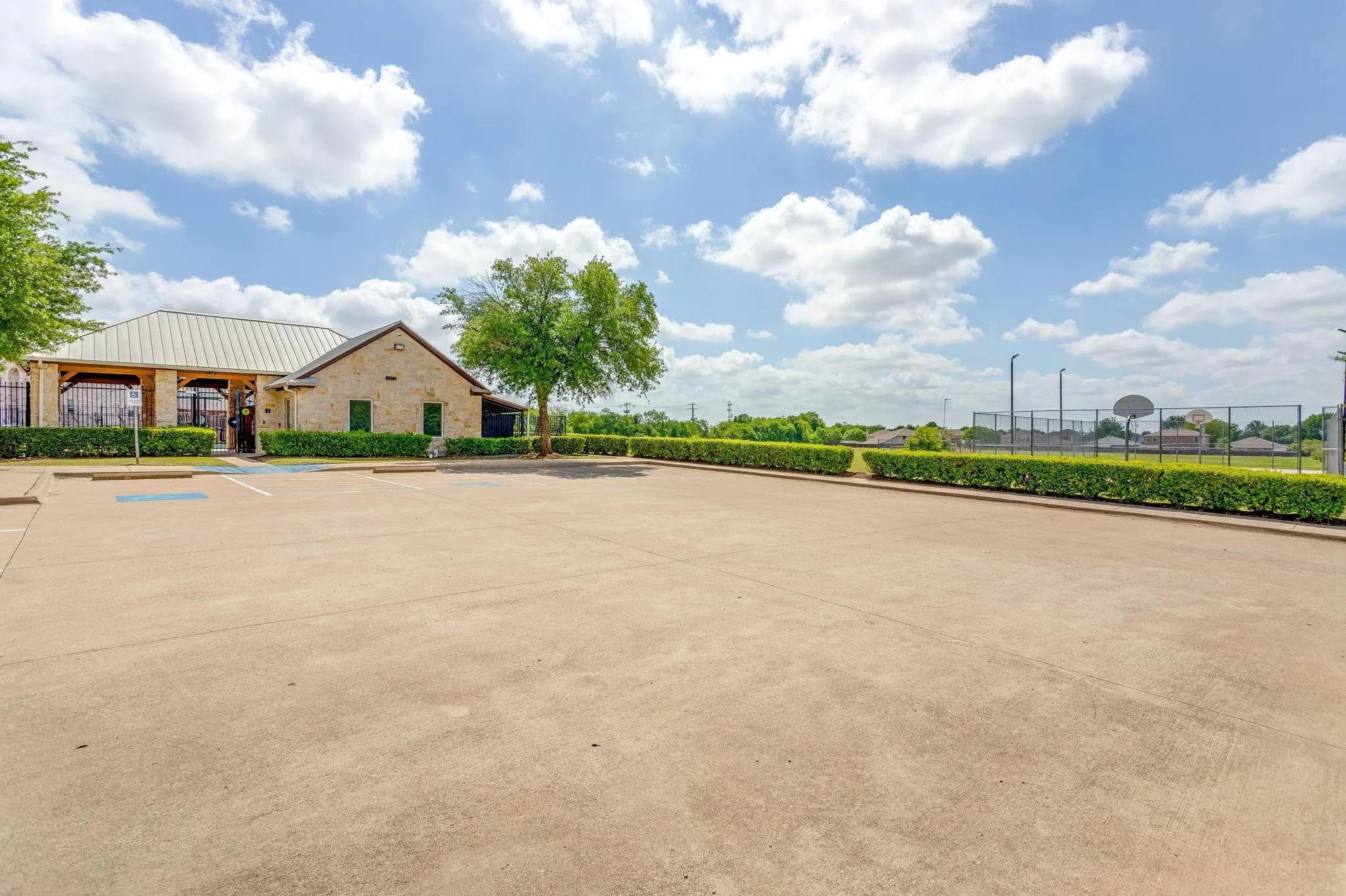 Stone-clad exterior facility featuring a metal roof, expansive paved parking area, manicured hedge landscaping, and a fenced court