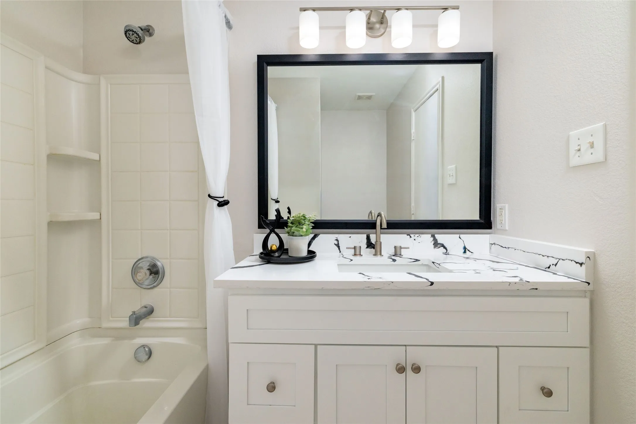 White vanity featuring a contrasting stone-finish countertop with an integrated rectangular sink and polished nickel hardware, complemented by a framed vanity mirror, four-bulb light fixture, and a tile-surround tub with built-in shelving