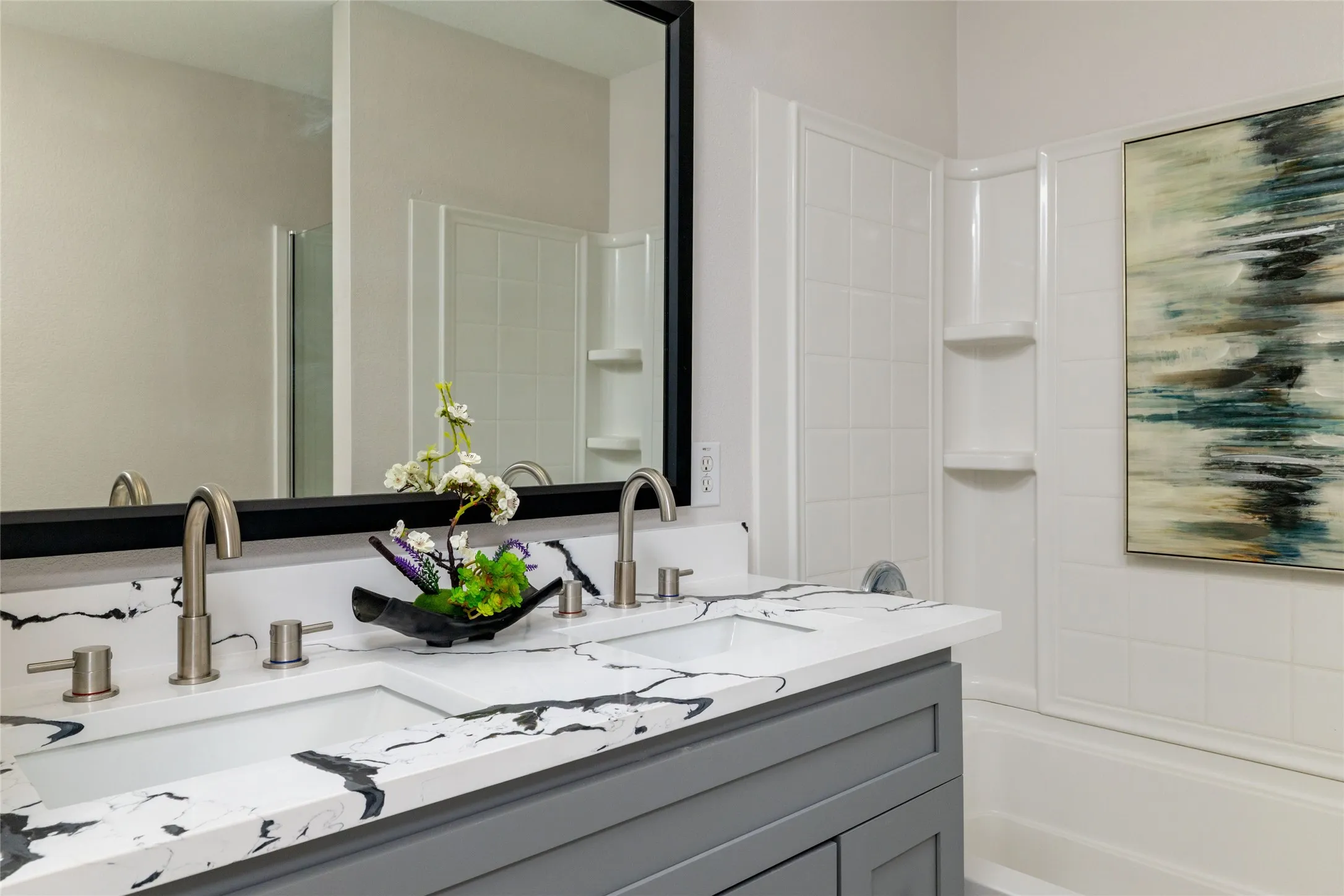 Double-sink vanity featuring a white surface with dark veining, grey shaker-style cabinetry, brushed nickel arched faucets, and a large rectangular mirror with a black frame