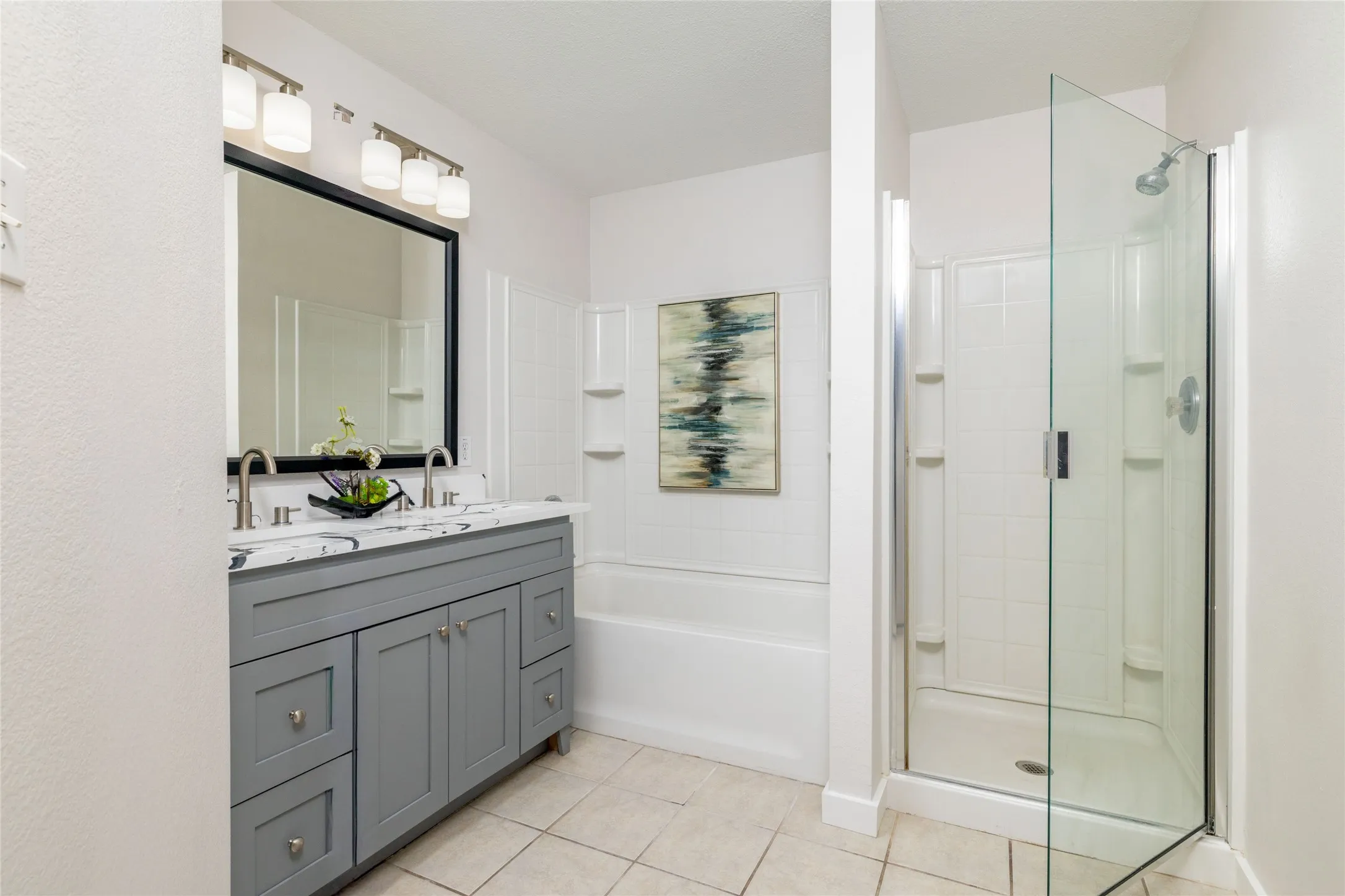 Bathroom featuring a dual-sink vanity with stone countertop, framed mirror with integrated vanity lighting, tiled tub-shower combination, and a separate glass-enclosed shower stall