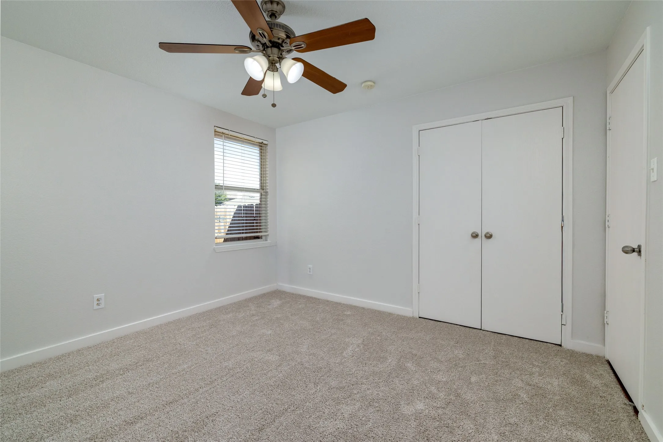 Neutral-toned wall-to-wall carpeting, ceiling fan with integrated lighting, single-pane window with horizontal blinds, and double-door closet