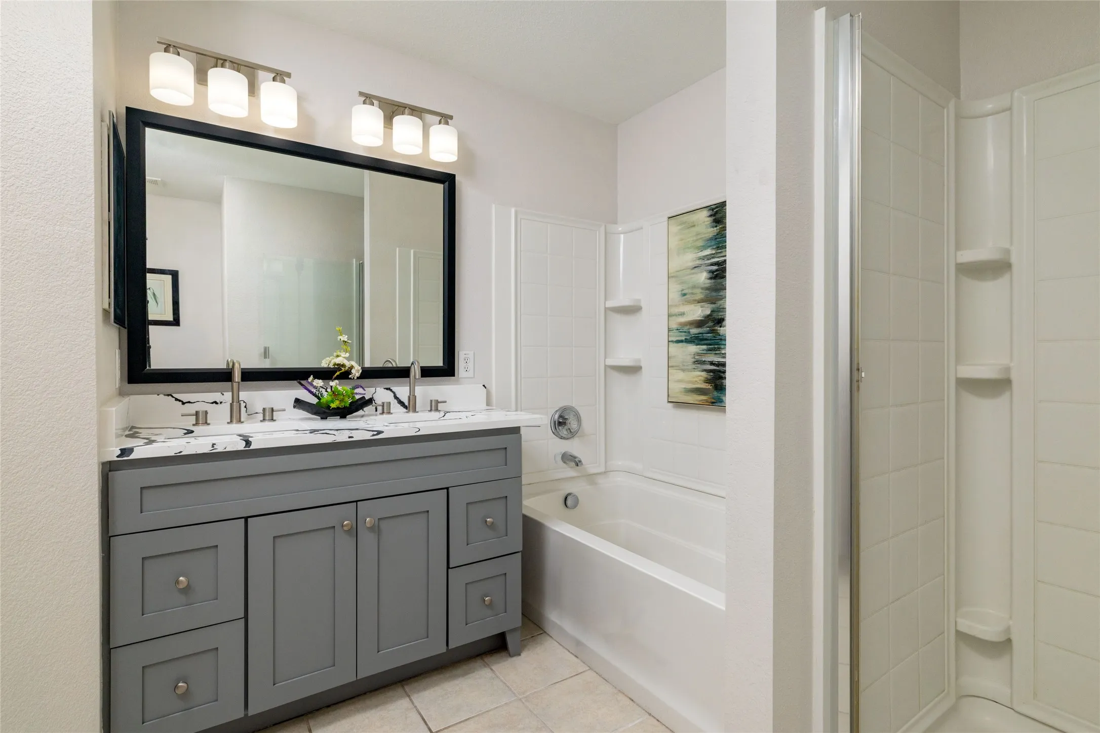 Dual-sink vanity with veined stone-finish countertop, Shaker-style cabinetry, framed vanity mirror, integrated bathtub-shower combination, and separate glass-enclosed shower stall