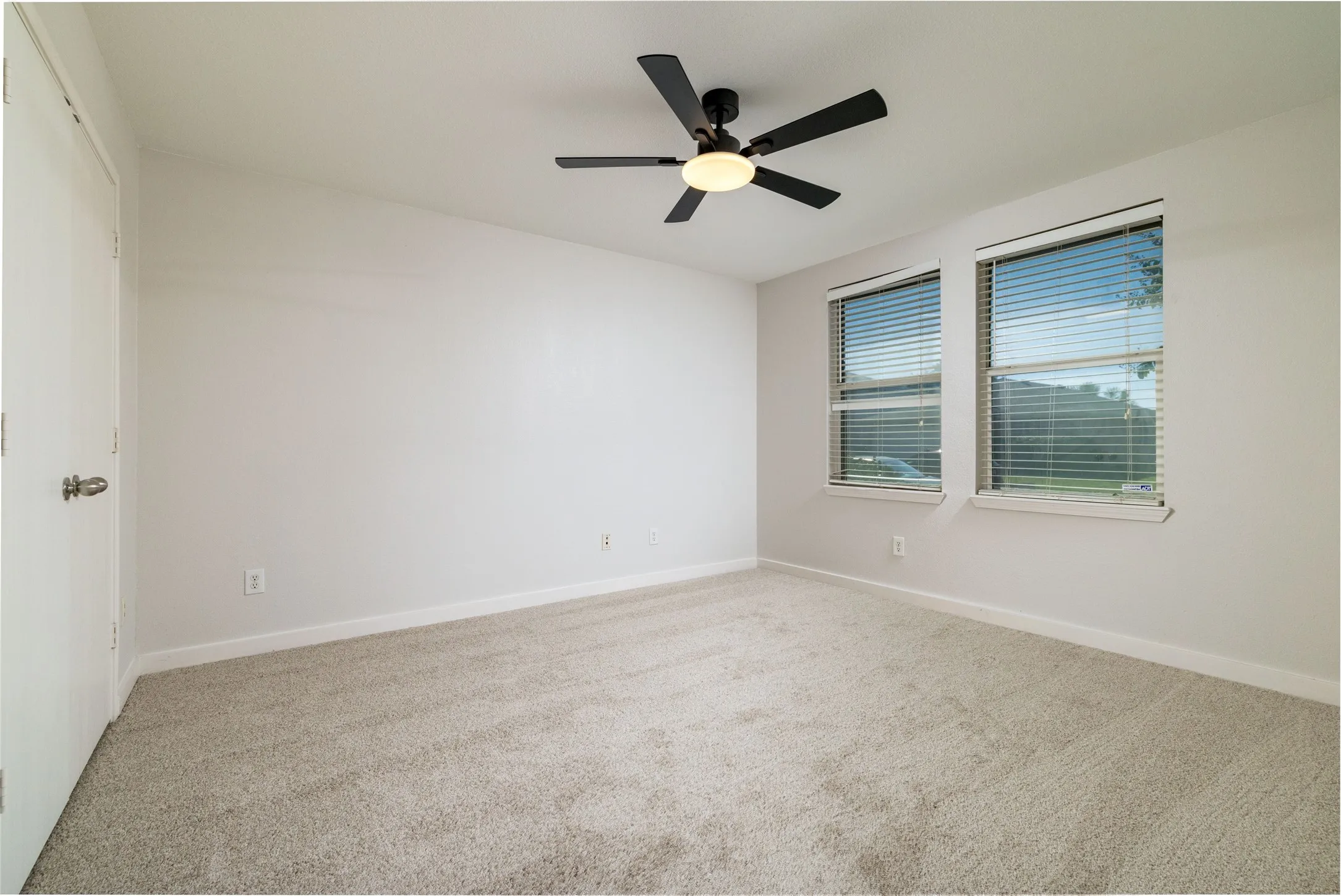 Interior space featuring neutral wall-to-wall carpeting, dual-pane windows with horizontal blinds, and a matte black ceiling fan with an integrated light fixture