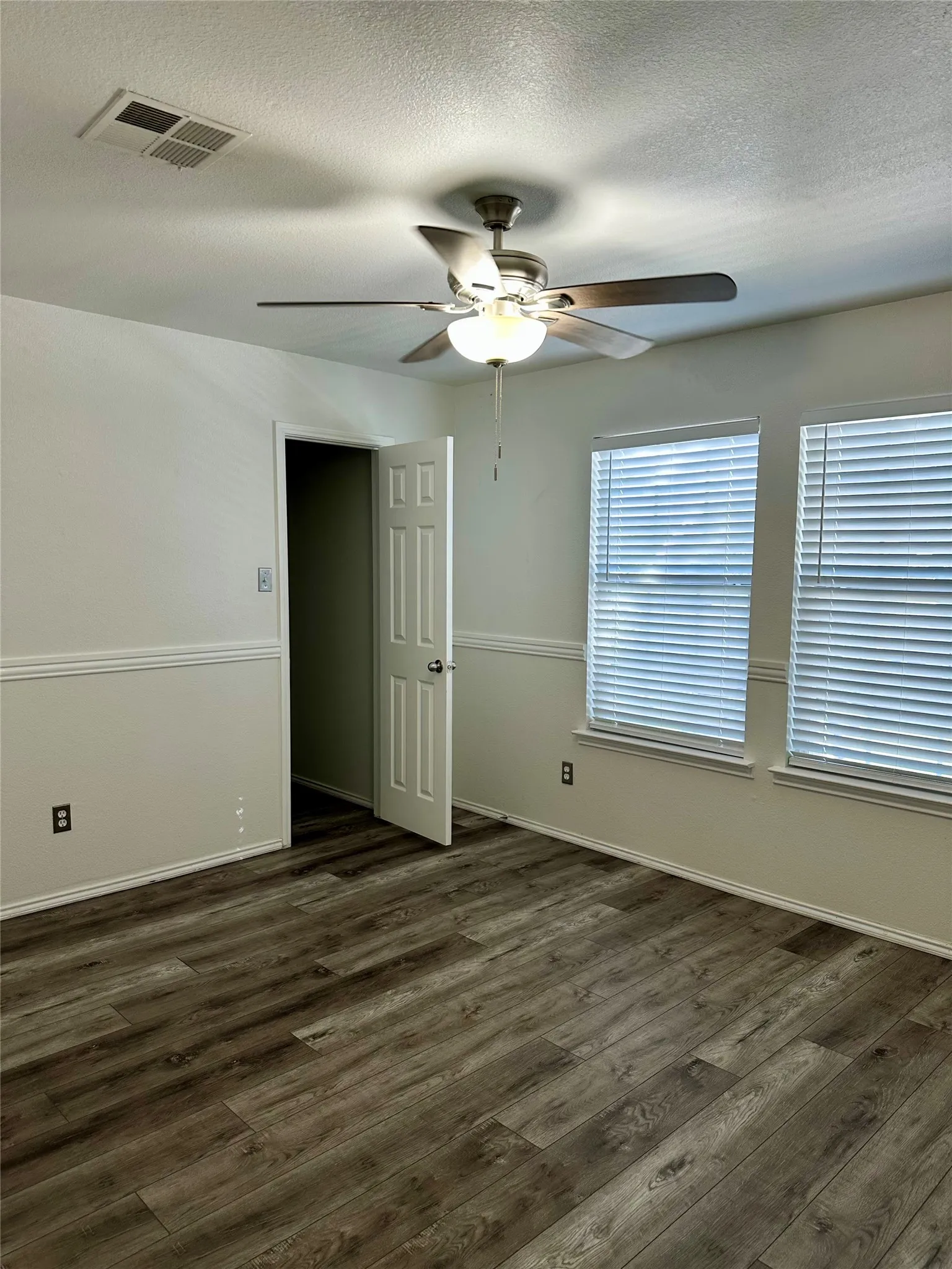 Interior room featuring wood-finish flooring, chair rail molding, dual windows with integrated blinds, and a brushed metal ceiling fan with light fixture