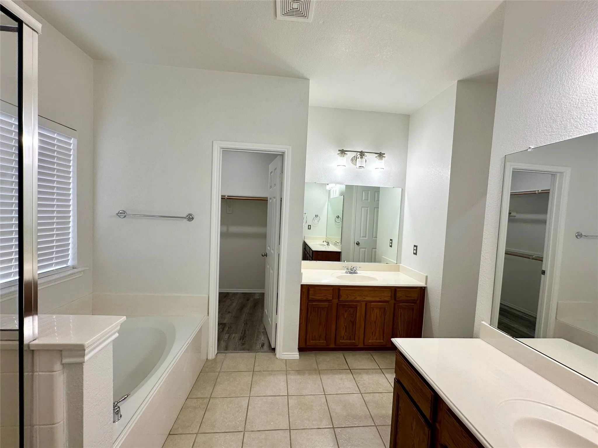 Bathroom featuring dual vanities with wood-finish cabinetry, solid-surface countertops, integrated sinks, and large wall-mounted mirrors, complemented by a soaking tub with tiled surround and ceramic floor tiling