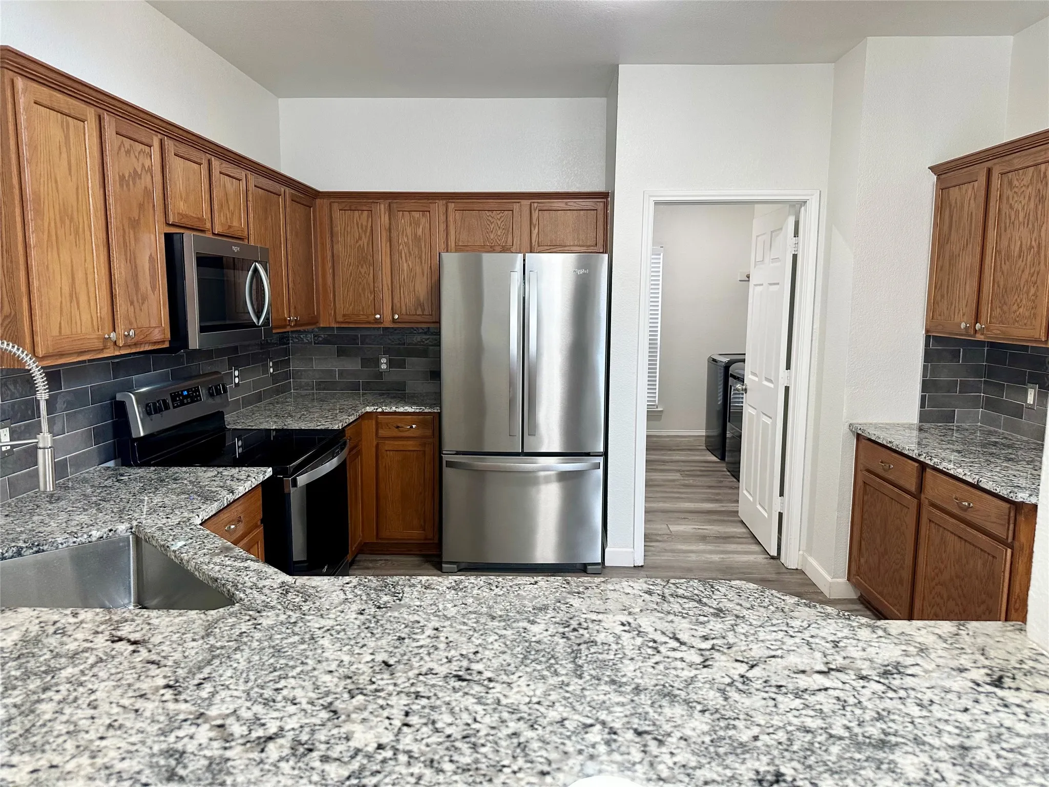 Kitchen featuring medium-tone wood cabinetry, stone countertops, dark tile backsplash, stainless steel appliances, and a stainless steel undermount sink