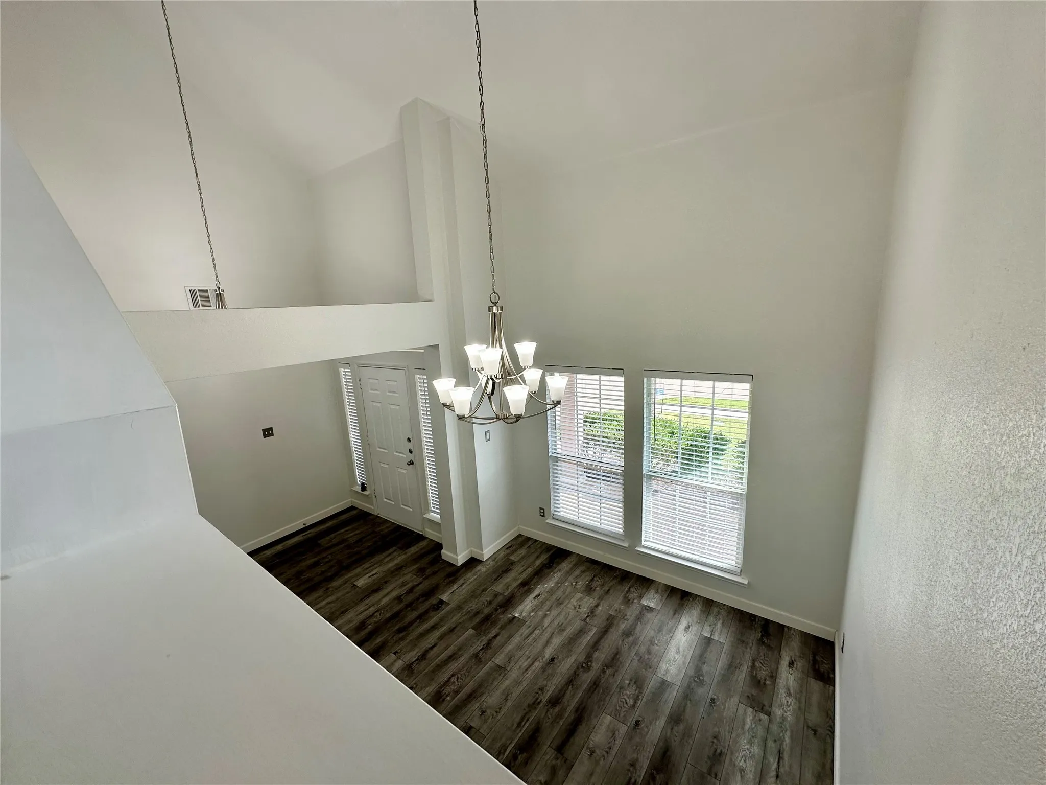 High-ceiling interior featuring wood-finish flooring, a multi-arm chandelier, and large windows with horizontal blinds