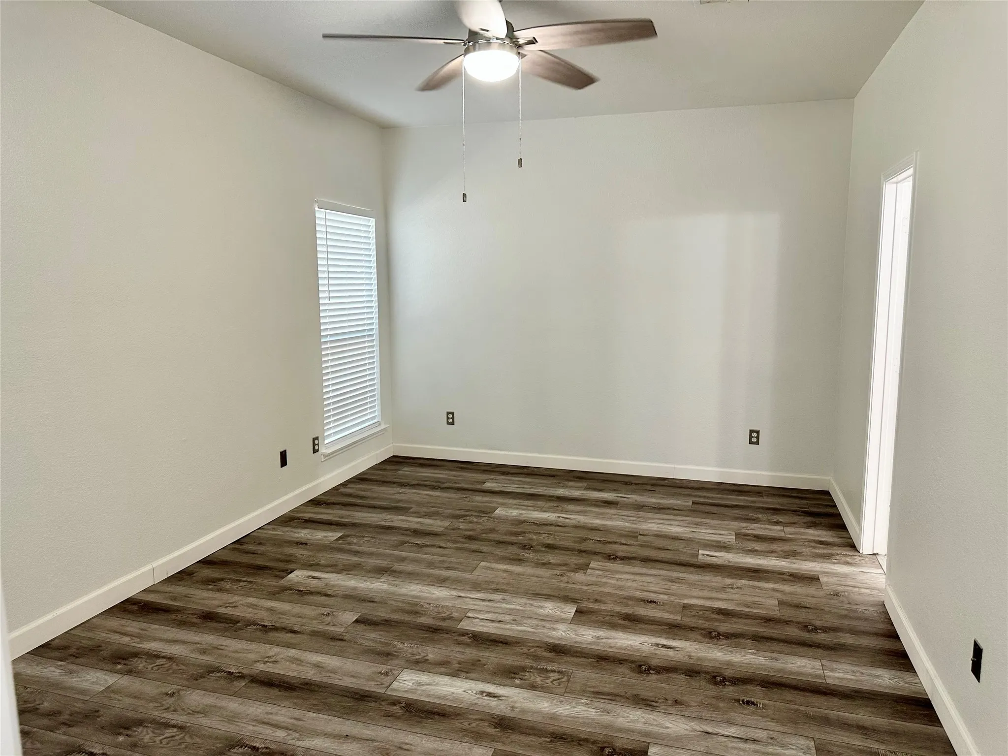 Neutral-toned interior space featuring wood-finish flooring, a ceiling fan with integrated light fixture, and horizontal blind window treatments