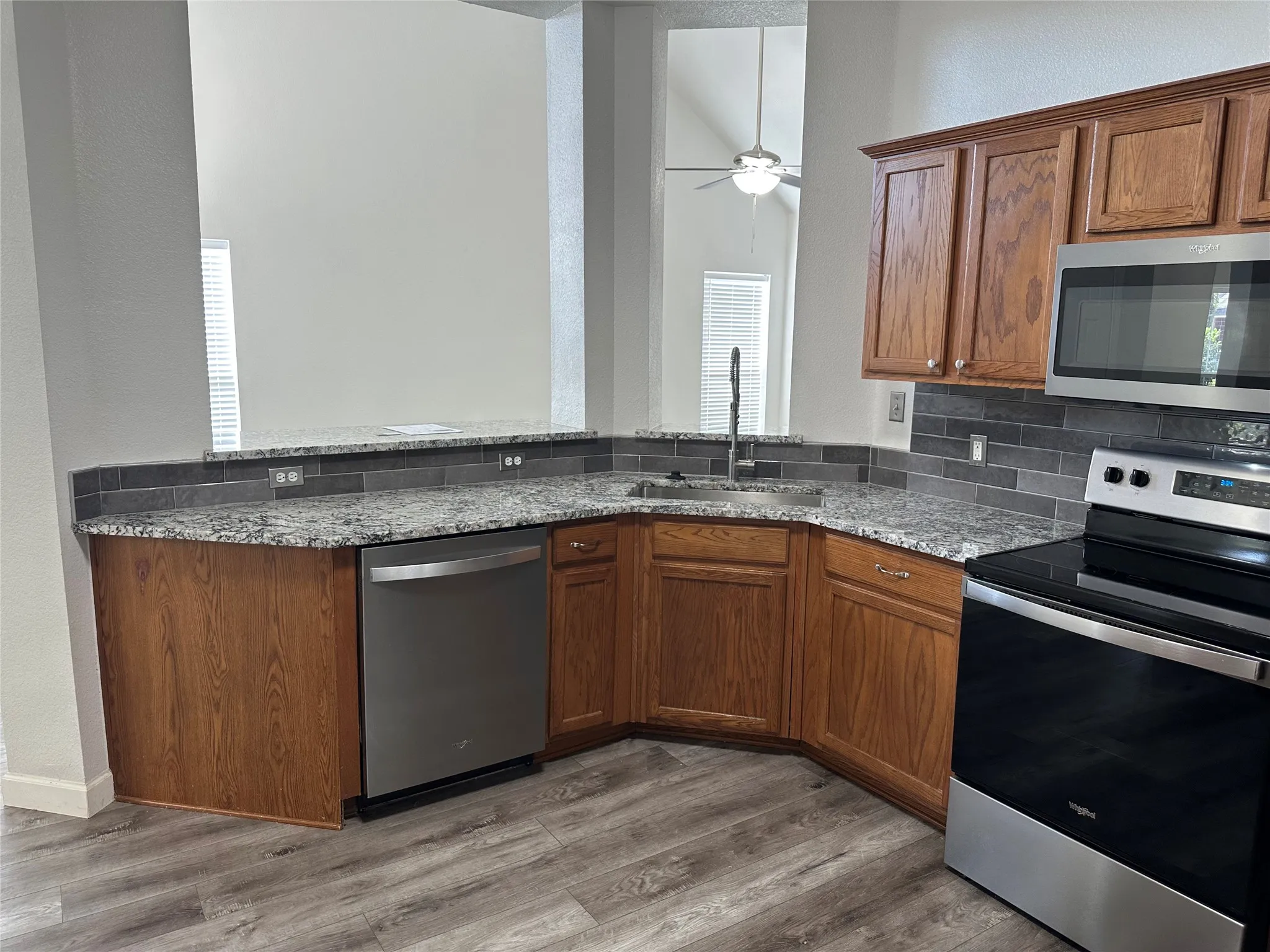 Kitchen featuring stone-finish countertops, stainless steel appliances, stained wood cabinetry, charcoal tile backsplash, and wood-finish flooring