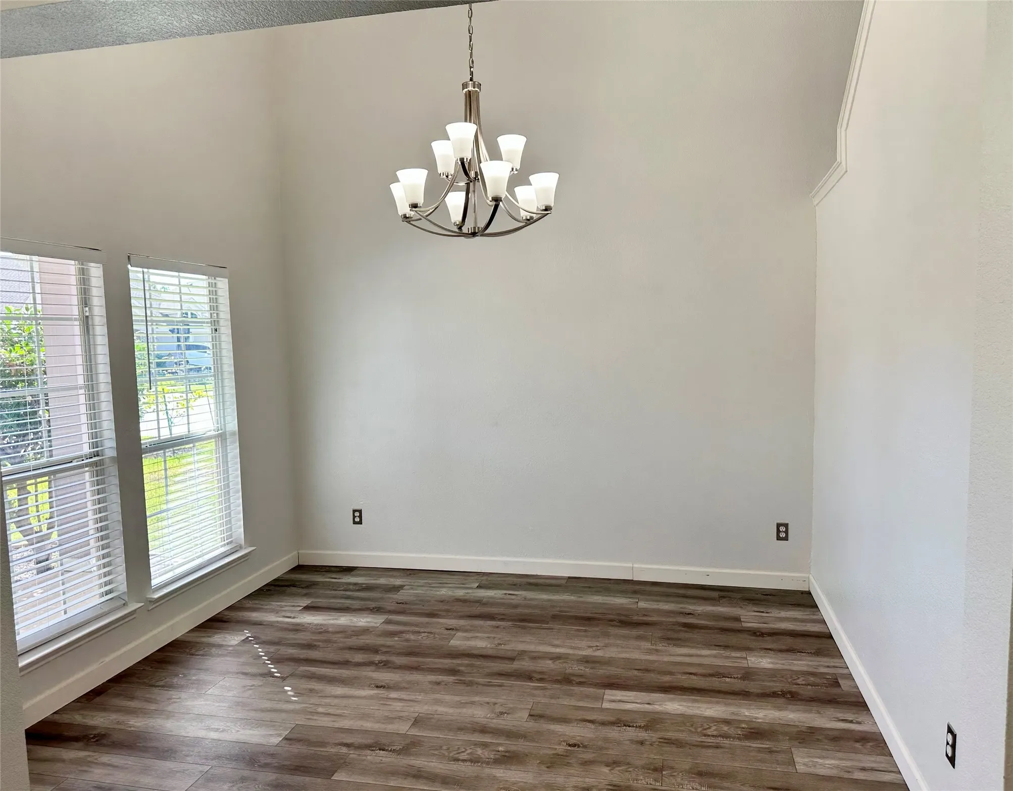 Vaulted ceiling, wood-finish flooring, multi-light brushed-nickel chandelier, and dual tall windows with horizontal blinds
