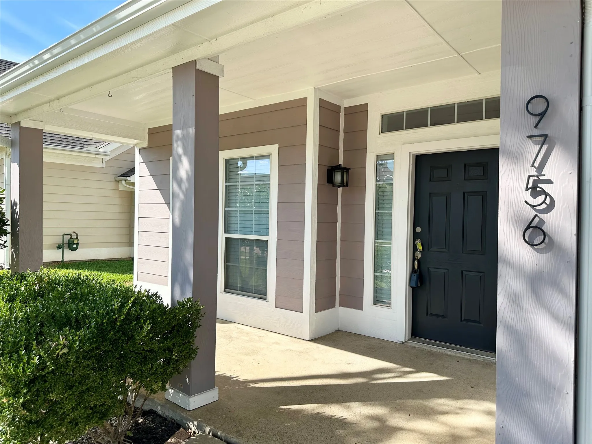 Covered entryway featuring horizontal lap siding, square support columns, a dark-toned solid core door with side light and transom window, and exterior wall-mounted lighting