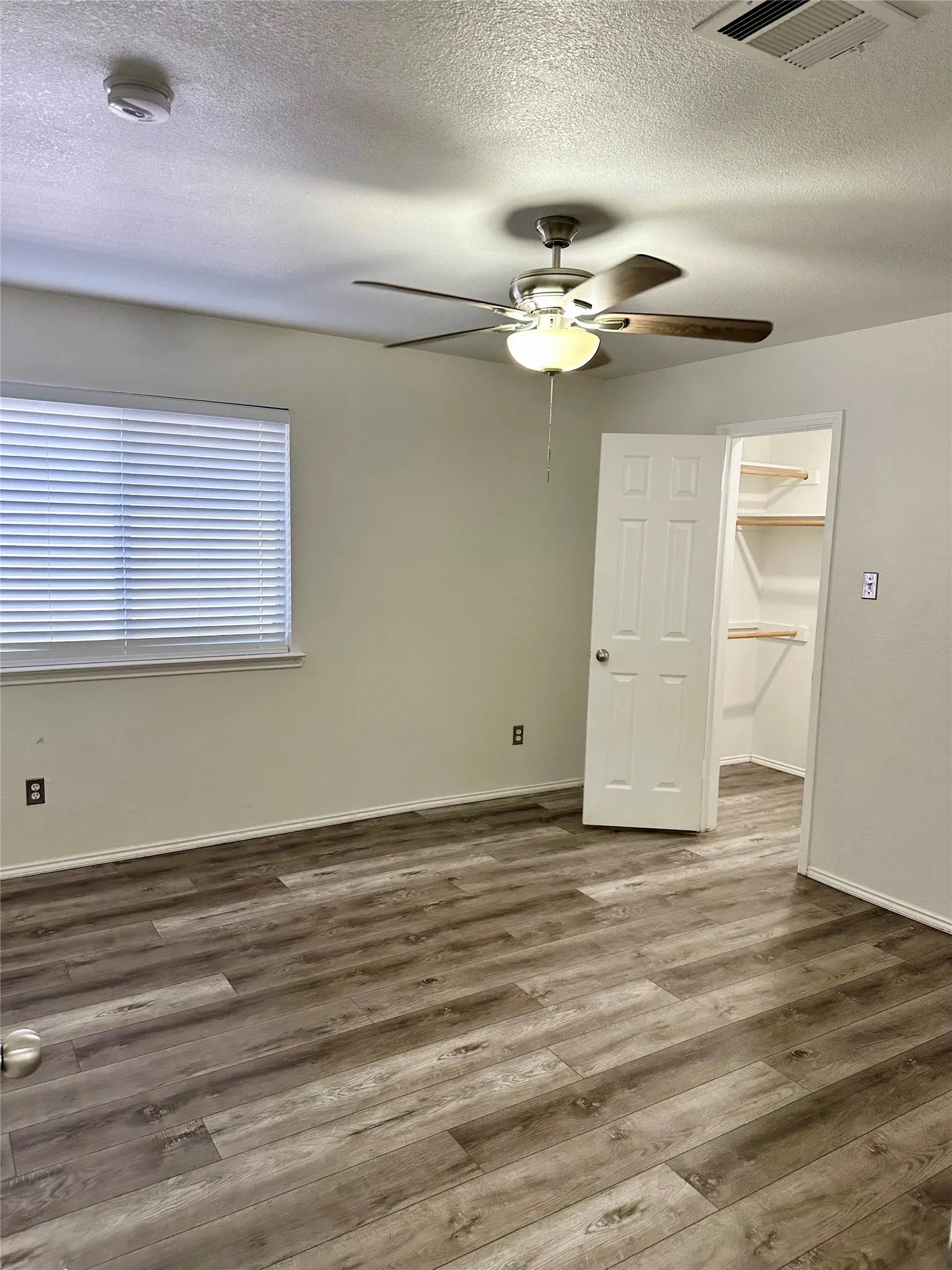 Wood-finish flooring, ceiling fan with integrated light fixture, window with horizontal blinds, and walk-in closet with built-in shelving