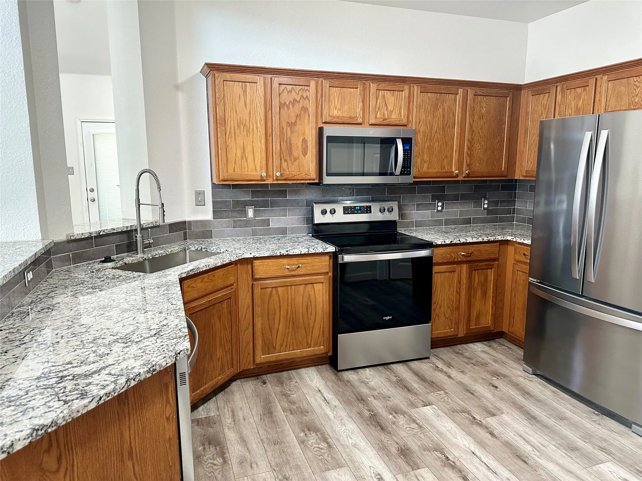 Kitchen featuring wood cabinetry, stone countertops, gray subway tile backsplash, stainless steel appliances, and wood-finish flooring