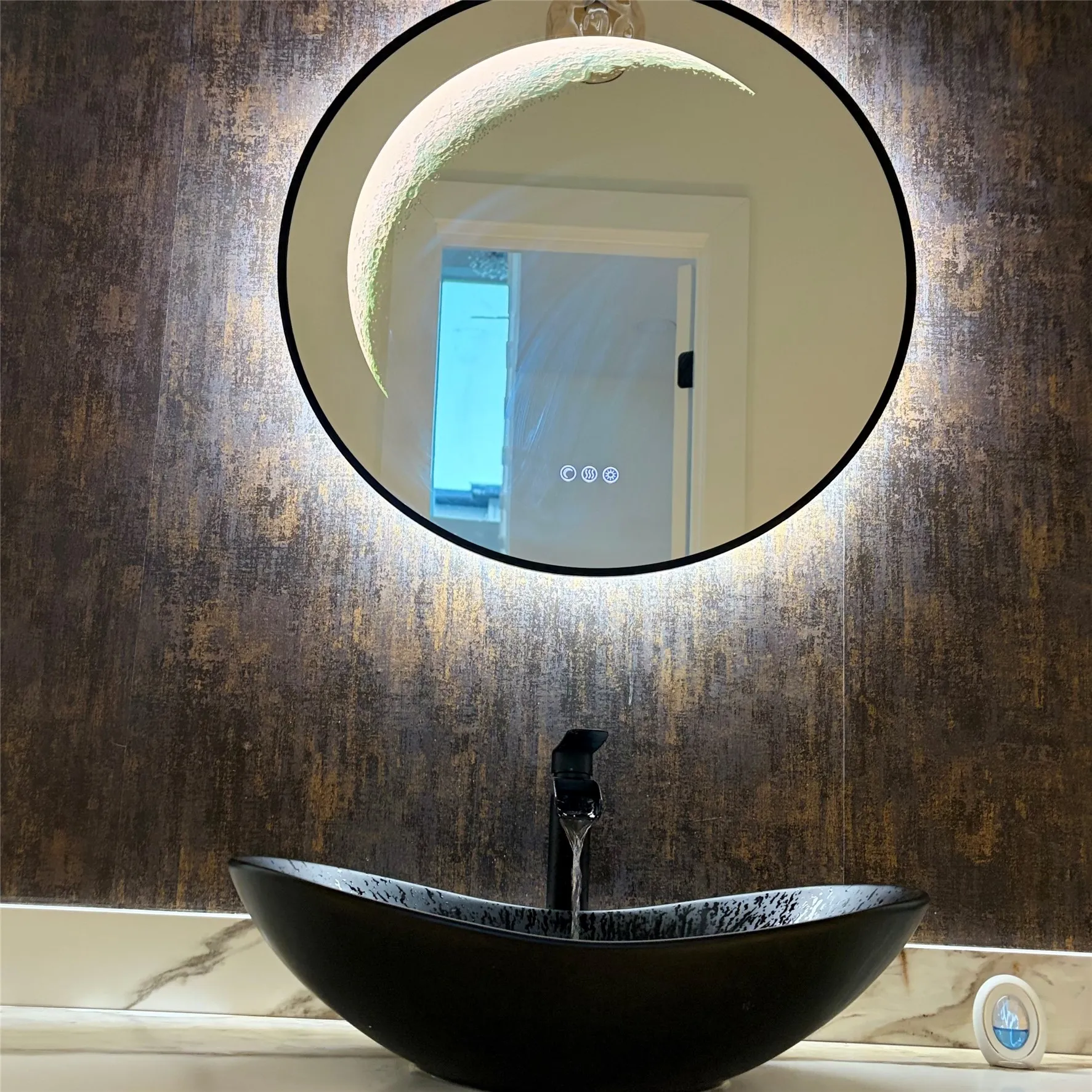 Round LED-backlit mirror featuring touch-control integration, matte black faucet, dark textured vessel sink, and stone-finish countertop with decorative backsplash