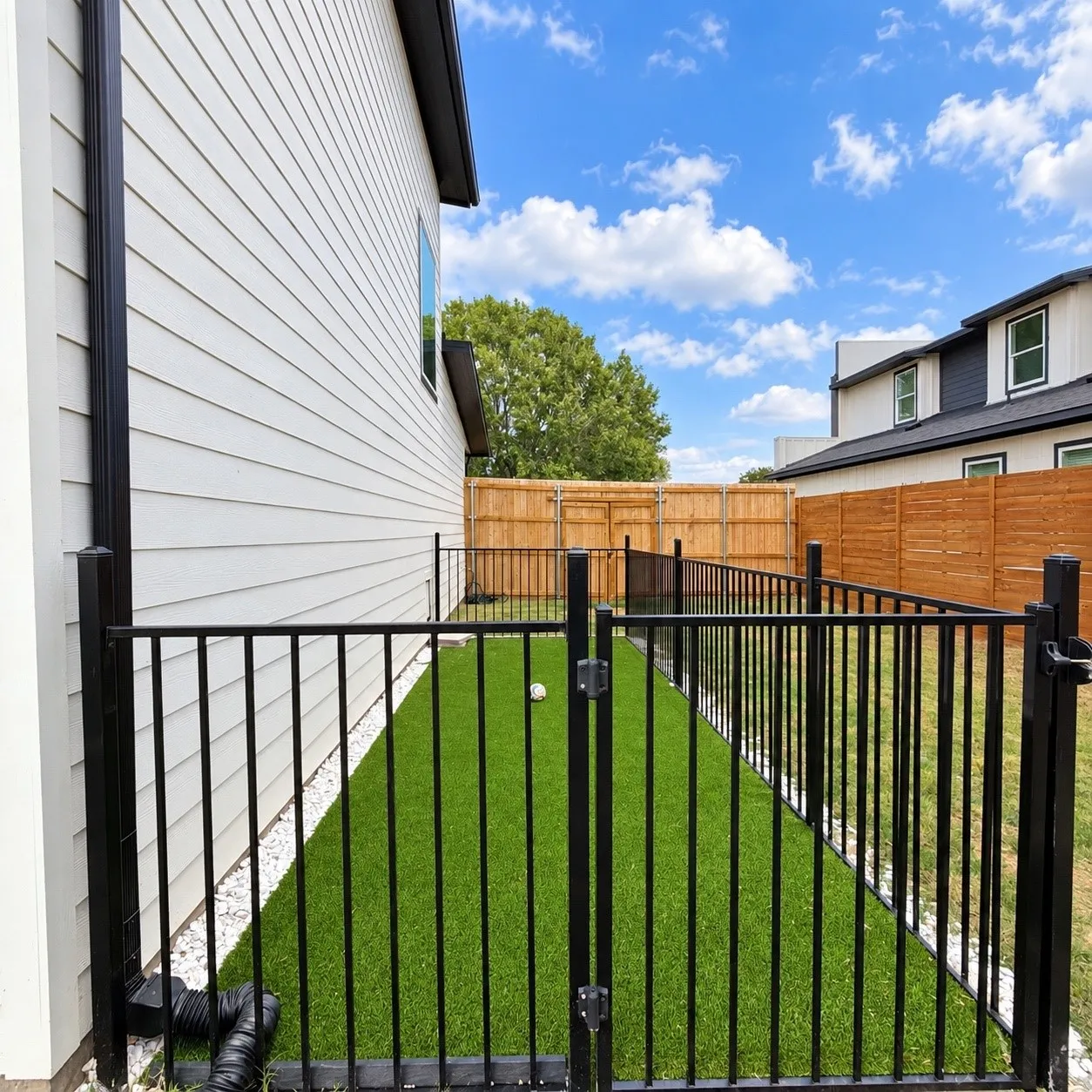 Side yard featuring synthetic turf, white stone border, black metal fencing, and wood privacy fencing