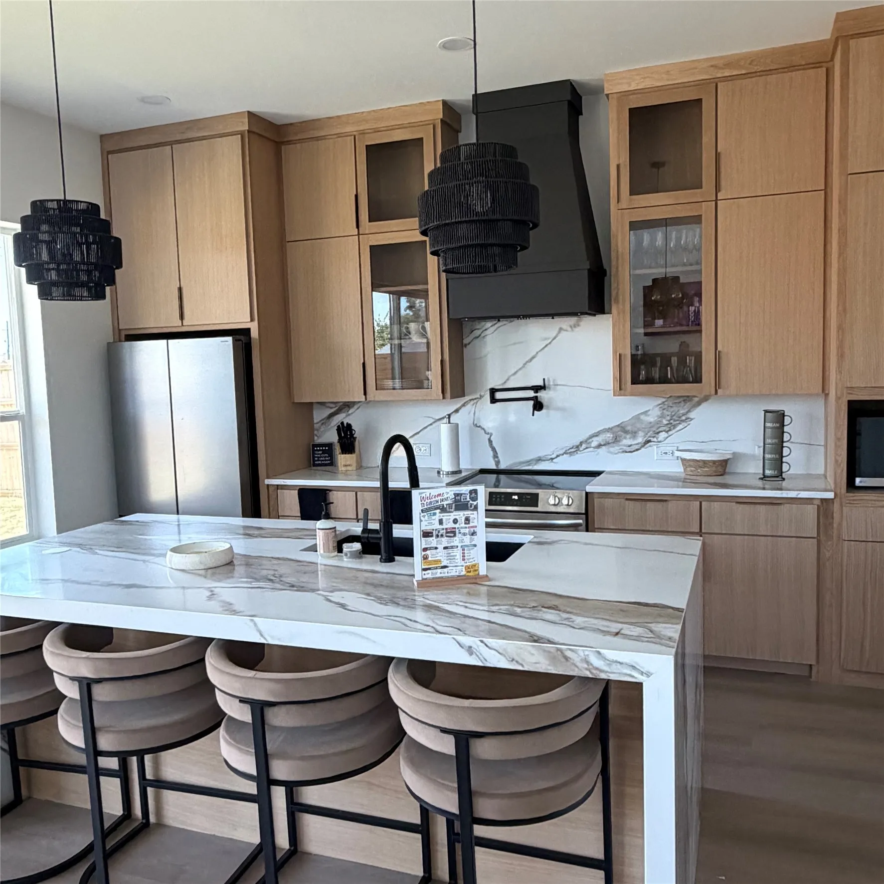 Kitchen featuring custom White Oak cabinetry, stone-patterned countertops with waterfall edge, full-height decorative backsplash, potfiller,black matte plumbing fixtures, and integrated stainless steel appliances