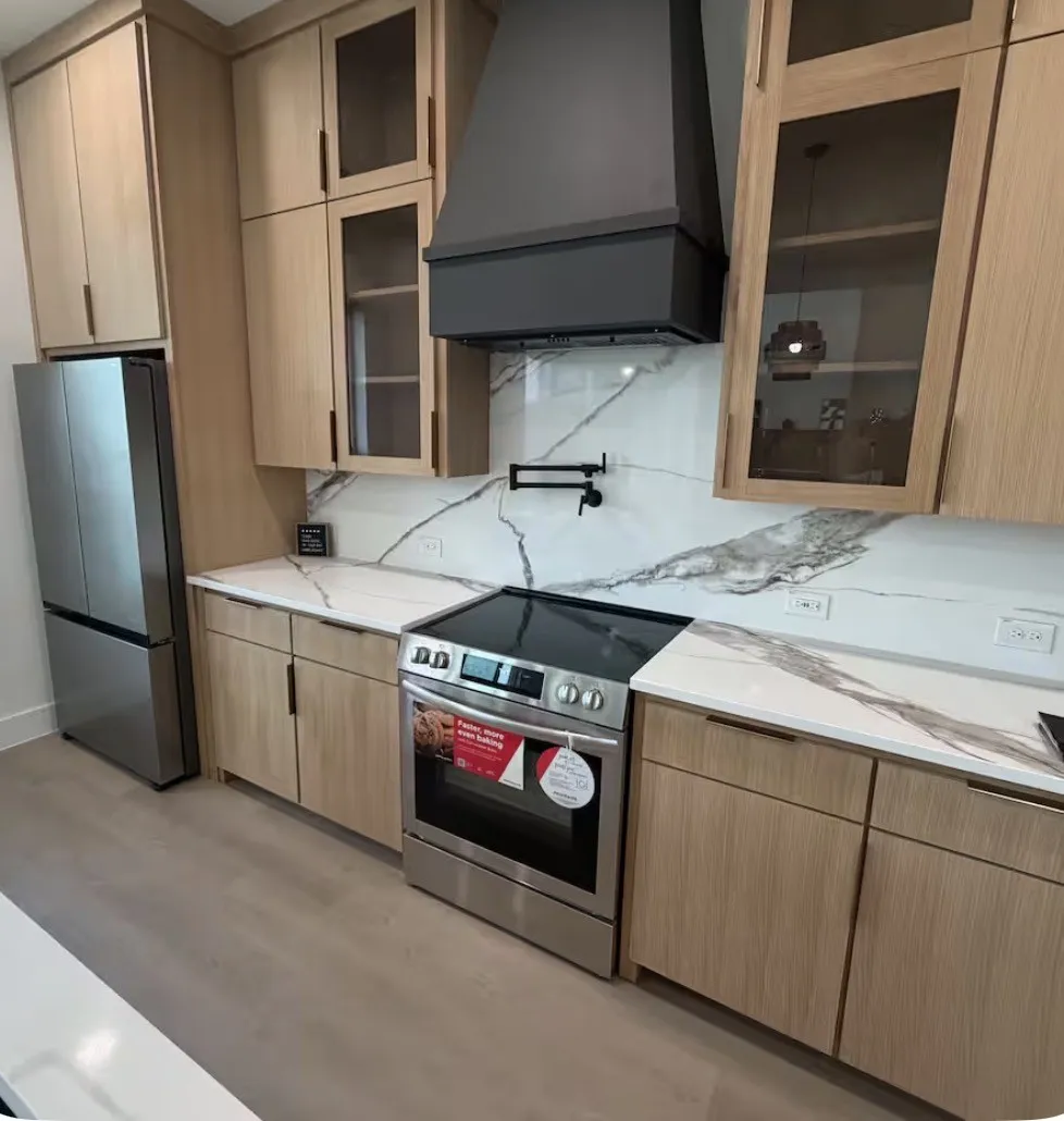 Modern kitchen featuring flat-panel wood-finish cabinetry, a full-height stone-finish backsplash, stainless steel appliances, a custom range hood, and an articulating wall-mounted pot filler