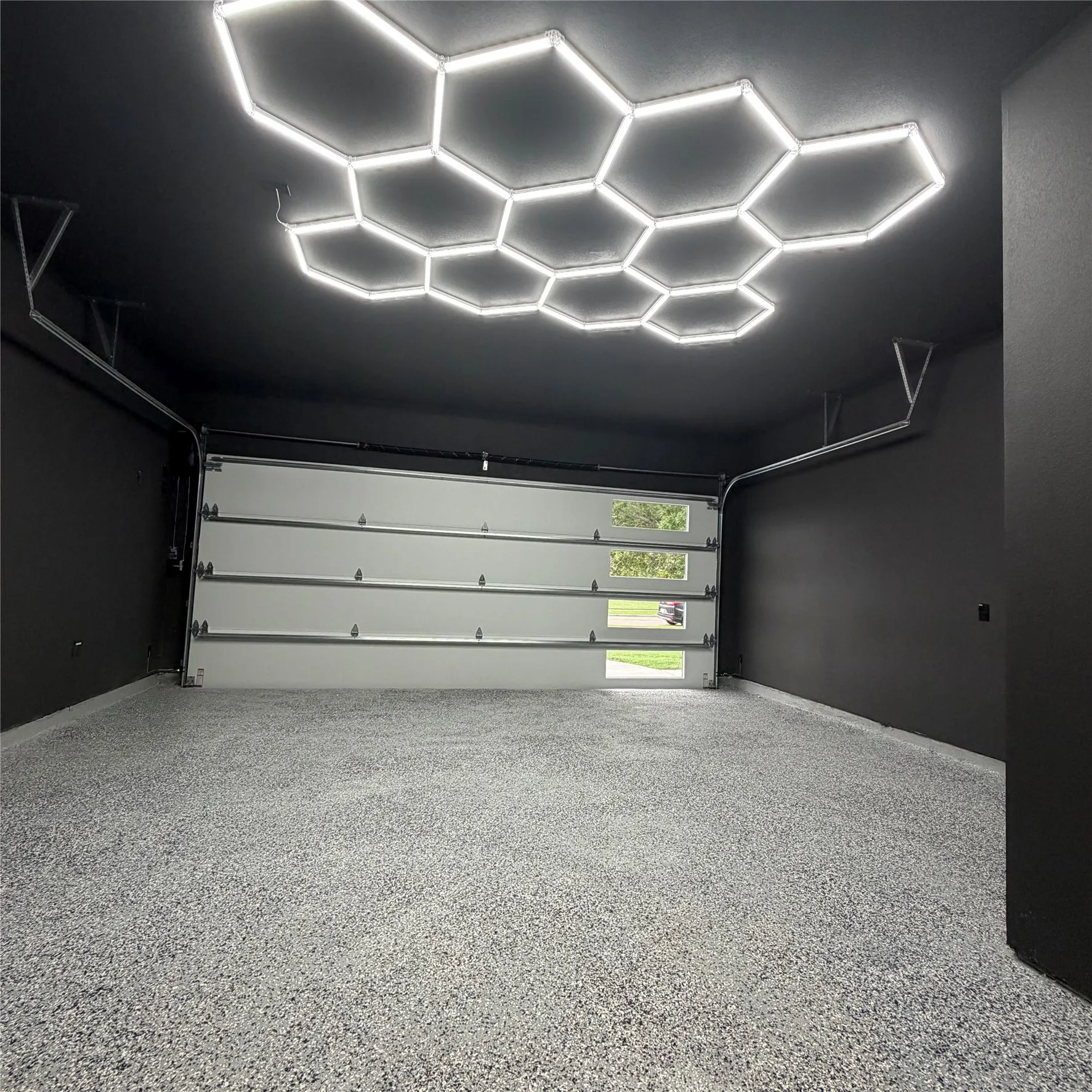 Garage interior featuring a custom honeycomb LED ceiling light array, high-contrast matte walls, speckled epoxy-coated flooring, and an insulated garage door with horizontal rectangular glass inserts