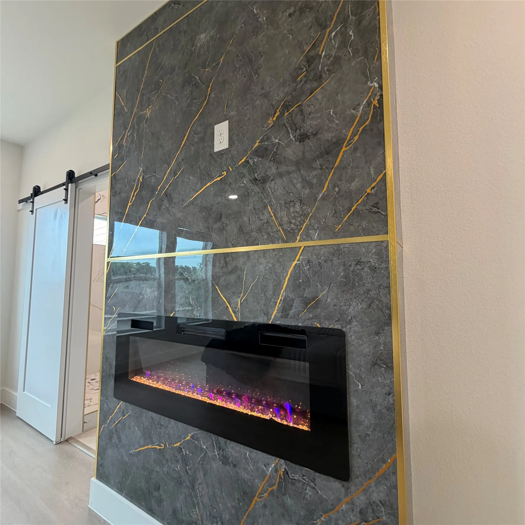 Floor-to-ceiling fireplace surround featuring dark stone-finish panels with gold-tone metal inlay, a built-in linear electric fireplace, and a white interior sliding barn door