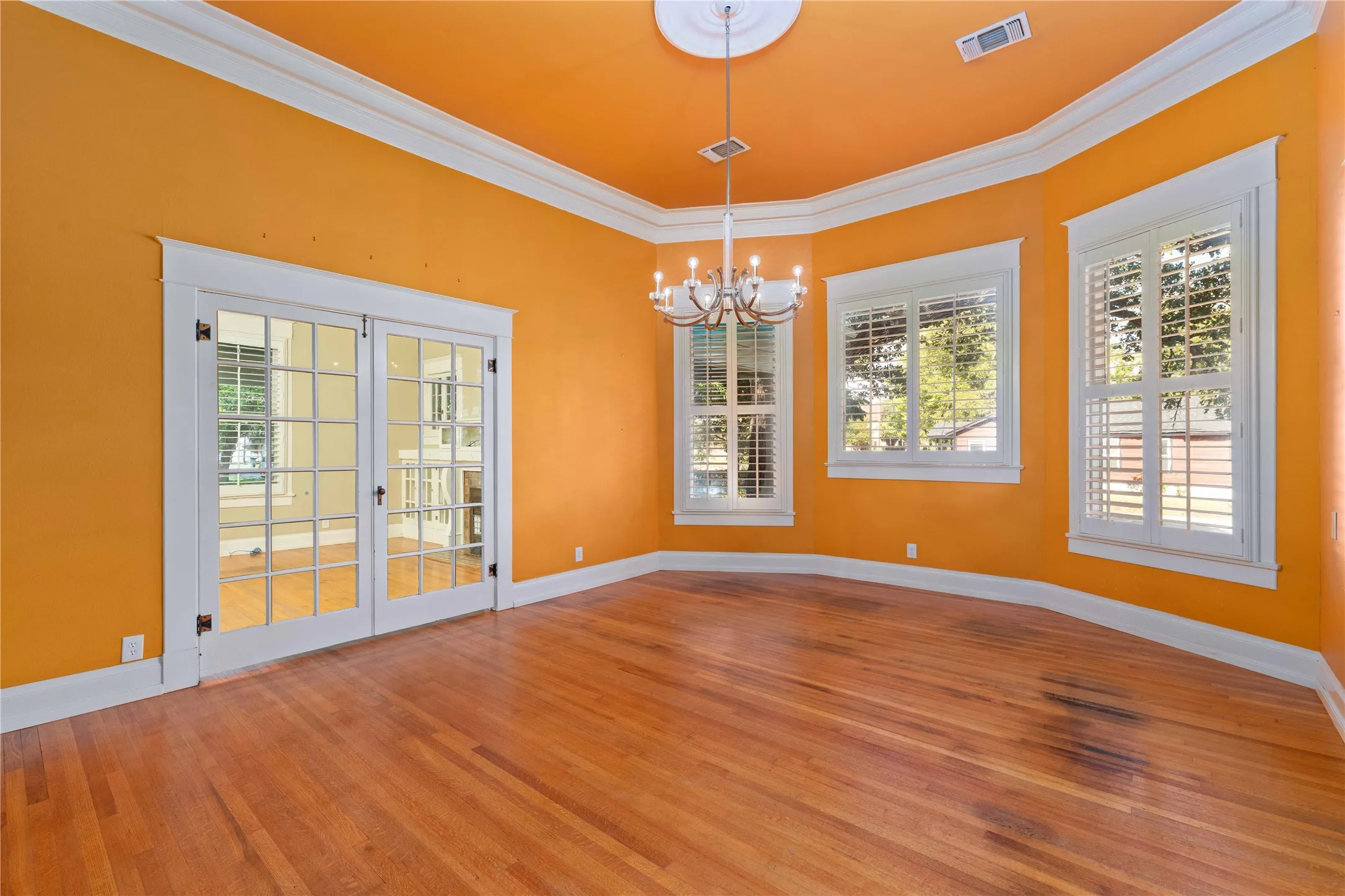 Room featuring wood-finish flooring, crown molding, double French doors, tiered chandelier with ceiling medallion, and white plantation shutters