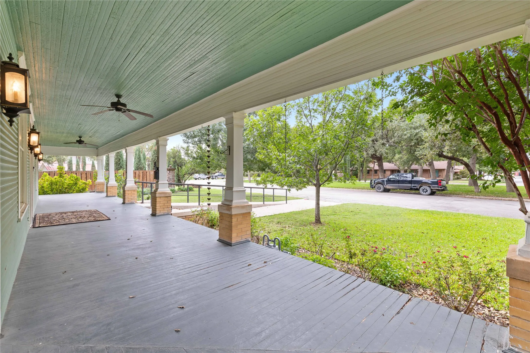 Covered wrap-around porch featuring wood-plank flooring, beadboard ceiling, brick-based columns, and wall-mounted exterior lighting