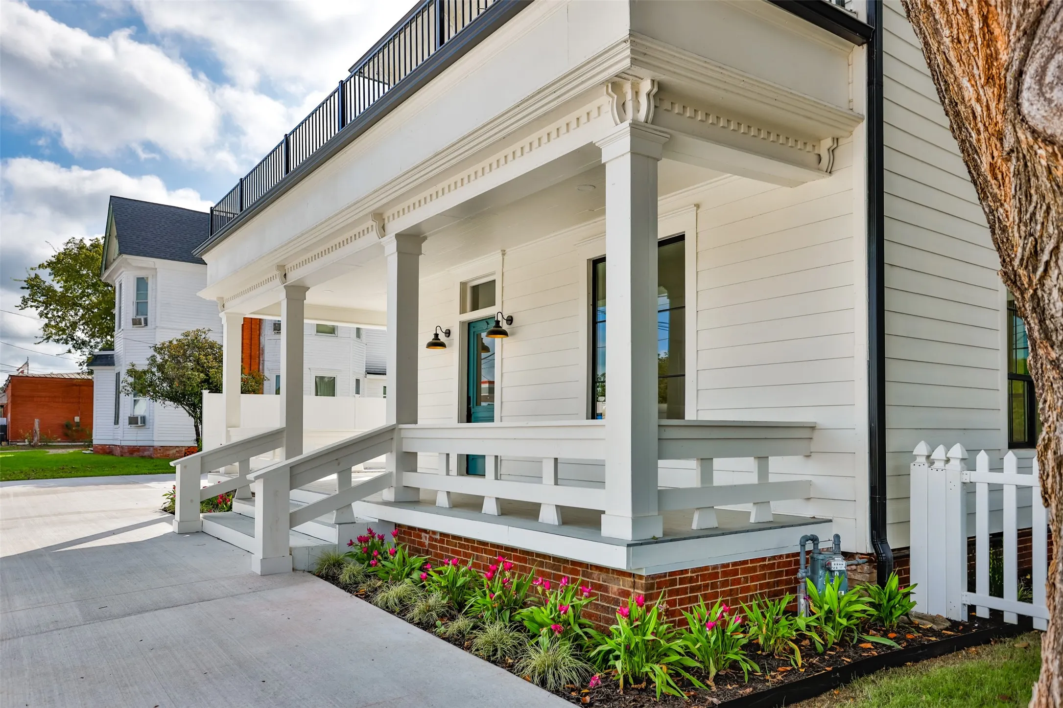 Covered front porch featuring white siding, decorative millwork columns, a brick foundation, and an upper-level balcony with metal railing