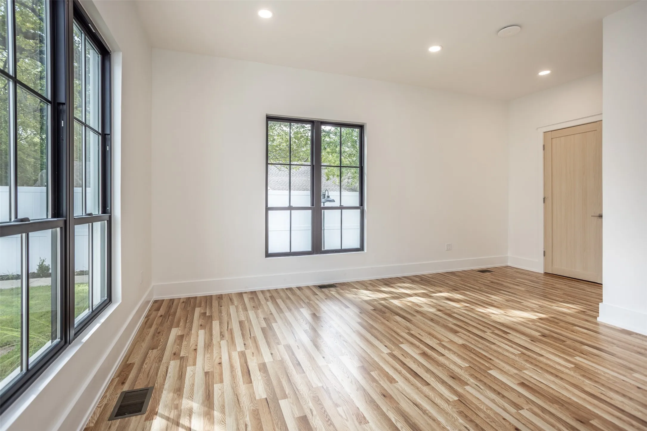 Light-filled room featuring wood-finish flooring, framed multi-pane windows, recessed ceiling lighting, and interior door