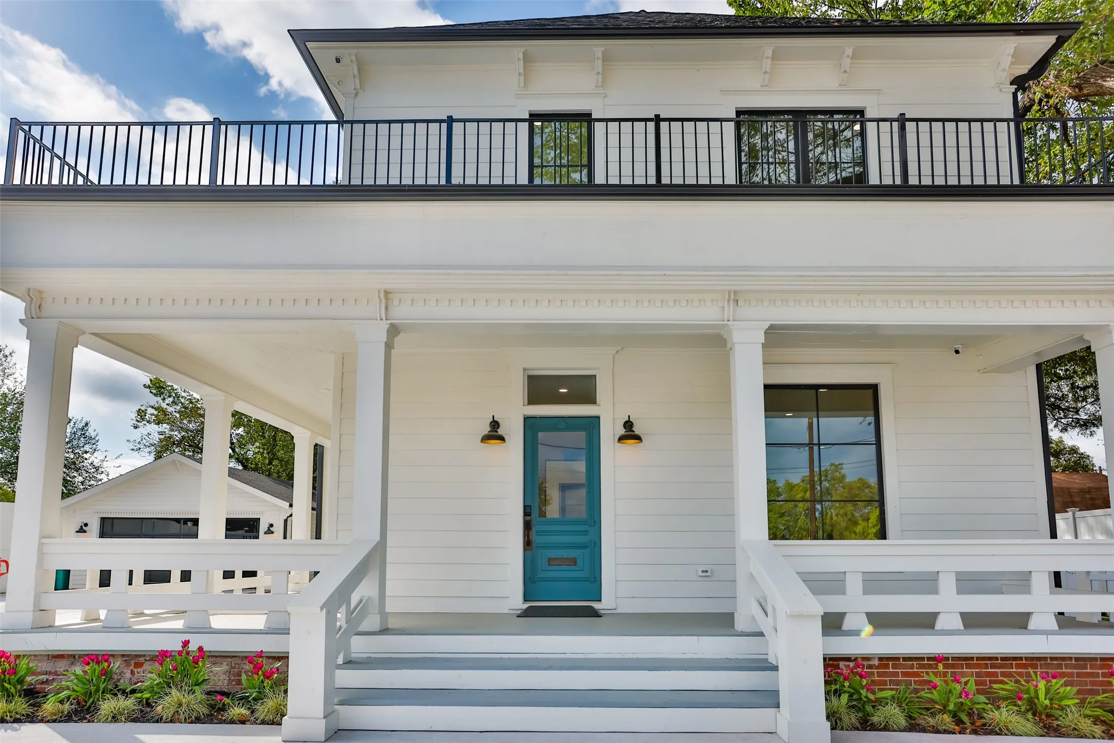 Two-story exterior featuring white siding, a covered front porch with a decorative railing, a second-story balcony with metal railings, and a blue painted front door