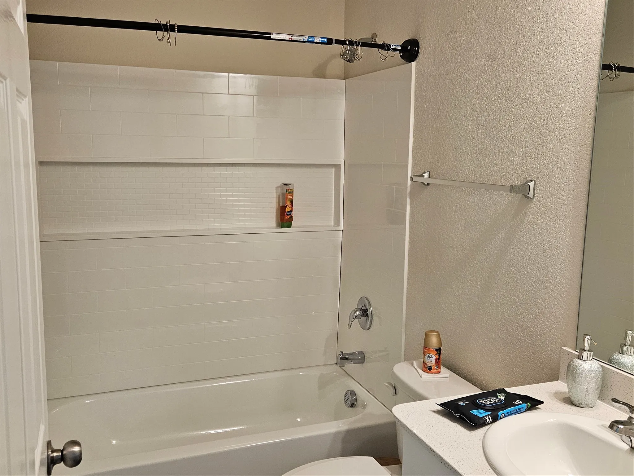 Full-width bathtub with white subway tile surround, integrated shower niche, light-toned countertop, and undermount vanity sink