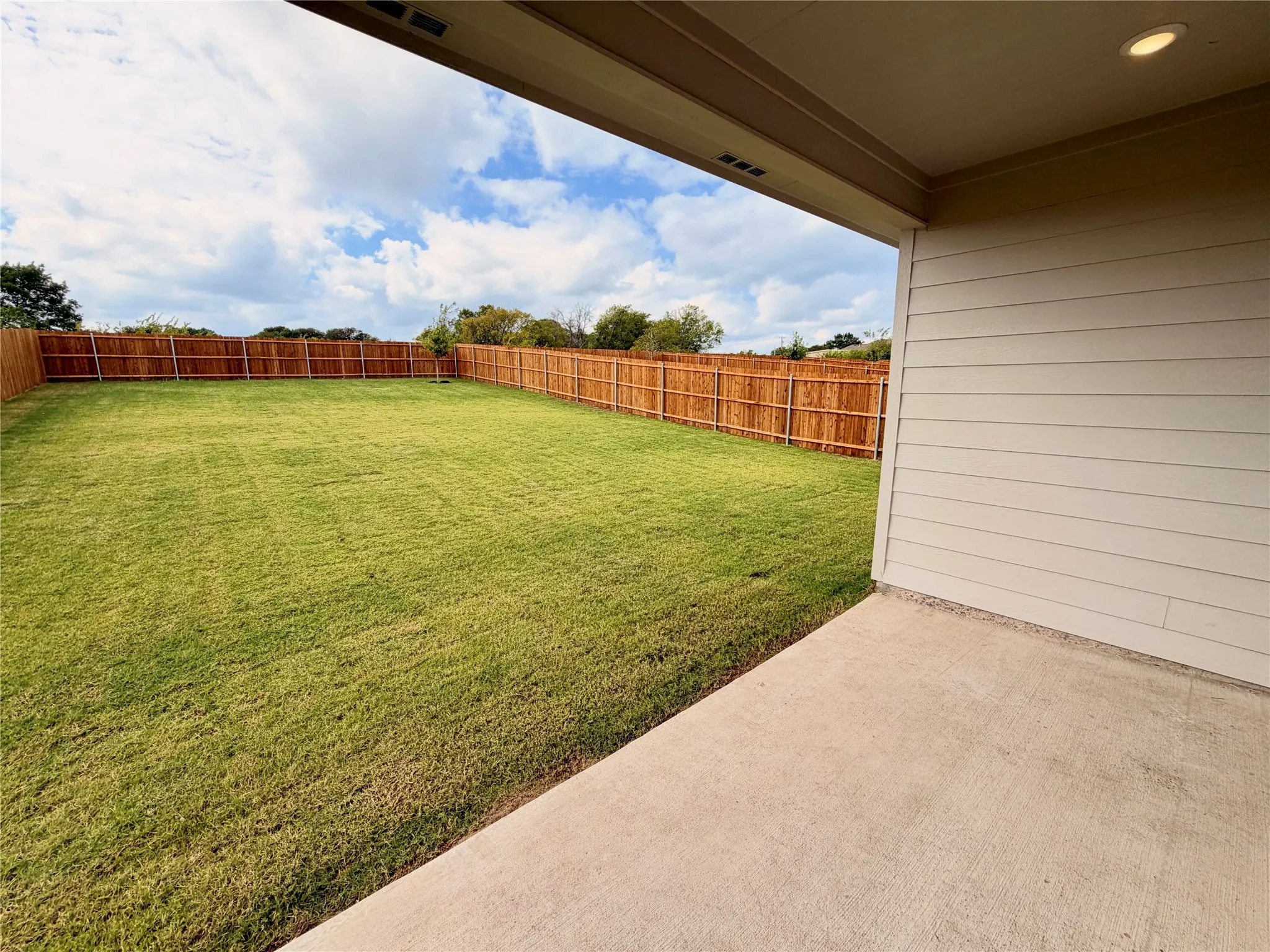 Covered concrete patio with horizontal siding exterior, expansive lawn, and wooden privacy fencing
