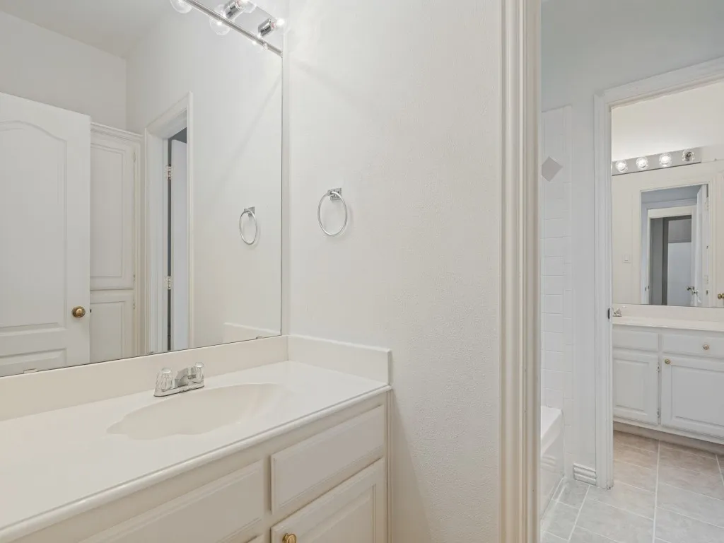White vanity cabinetry with a solid surface countertop, integrated sink, full-width vanity mirror, and tile flooring