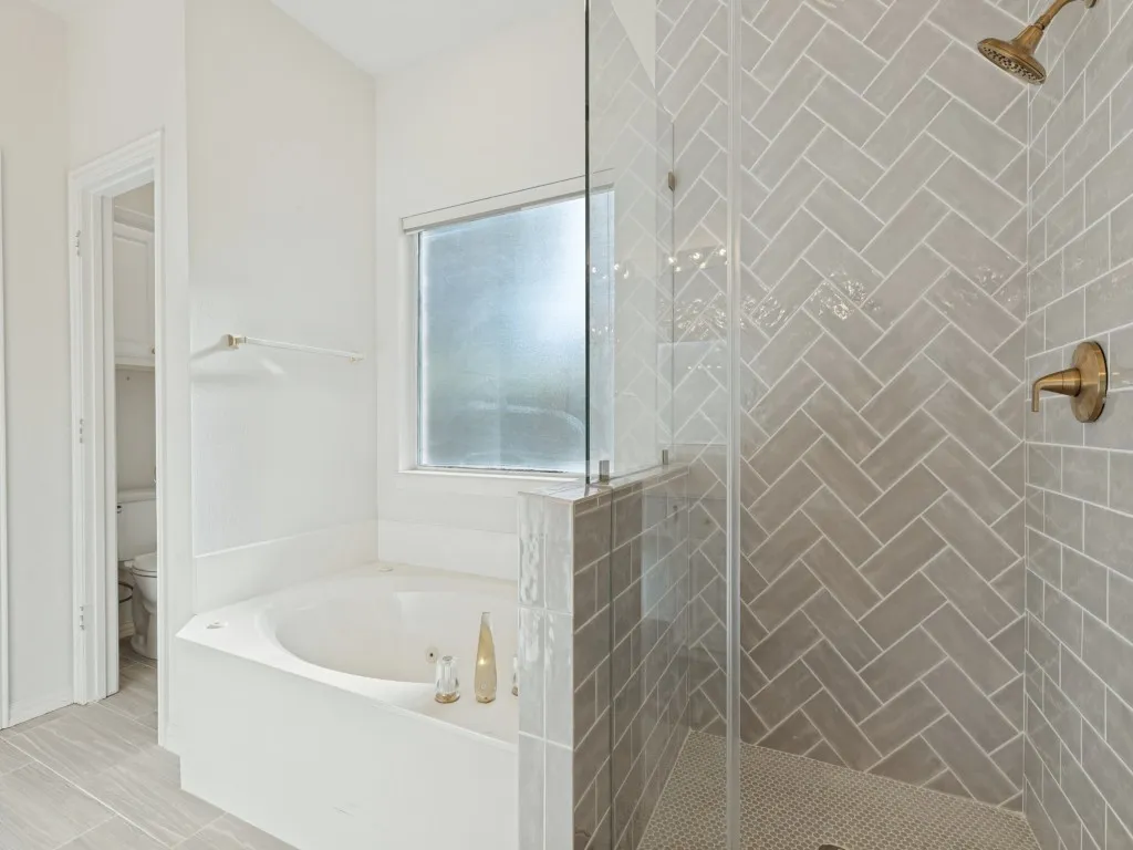 Shower enclosure featuring herringbone-patterned wall tile, frameless glass door, and metallic finish fixtures, complemented by a built-in soaking tub